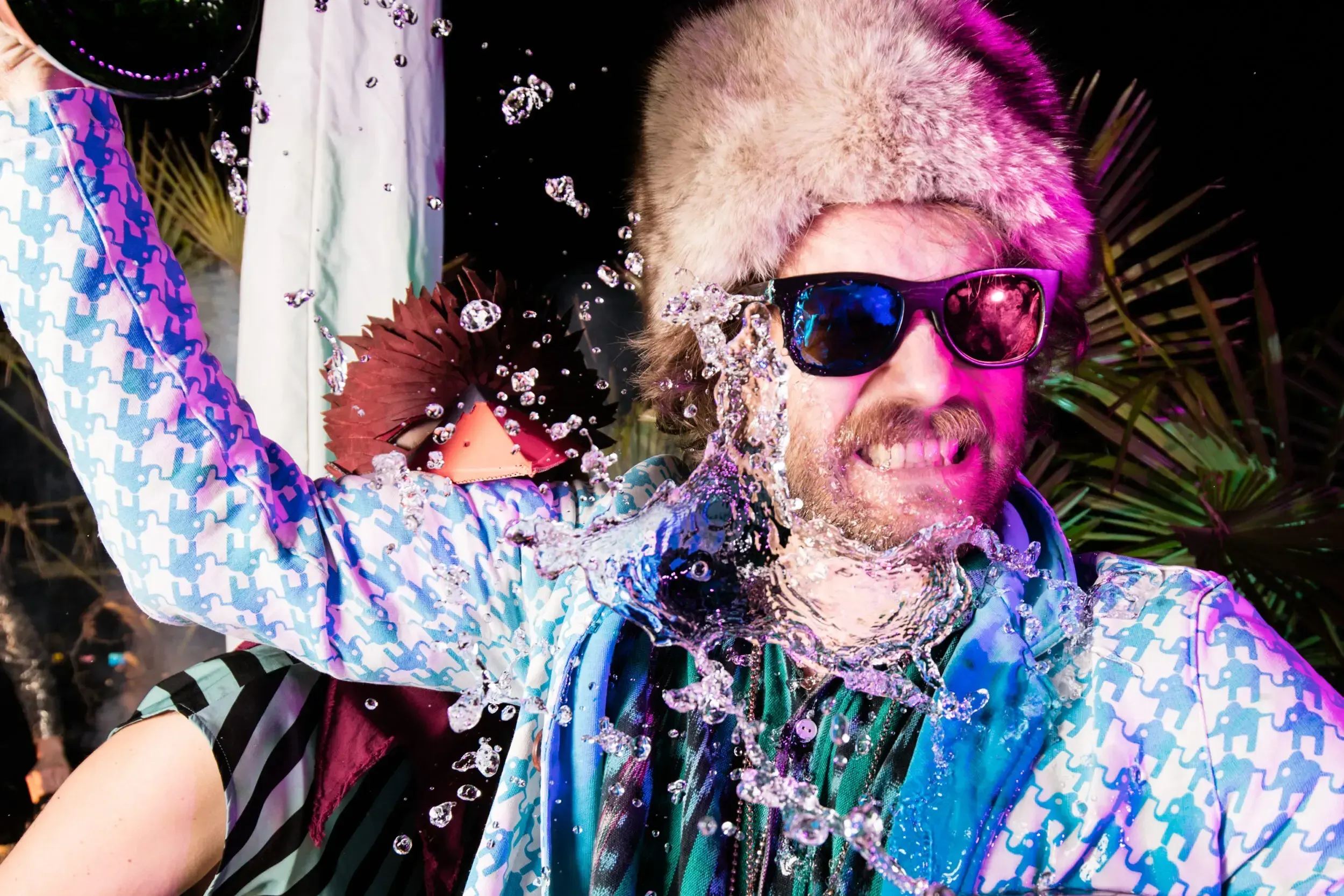 Guest in a fur hat and sunglasses with water splashing across his face at the party.