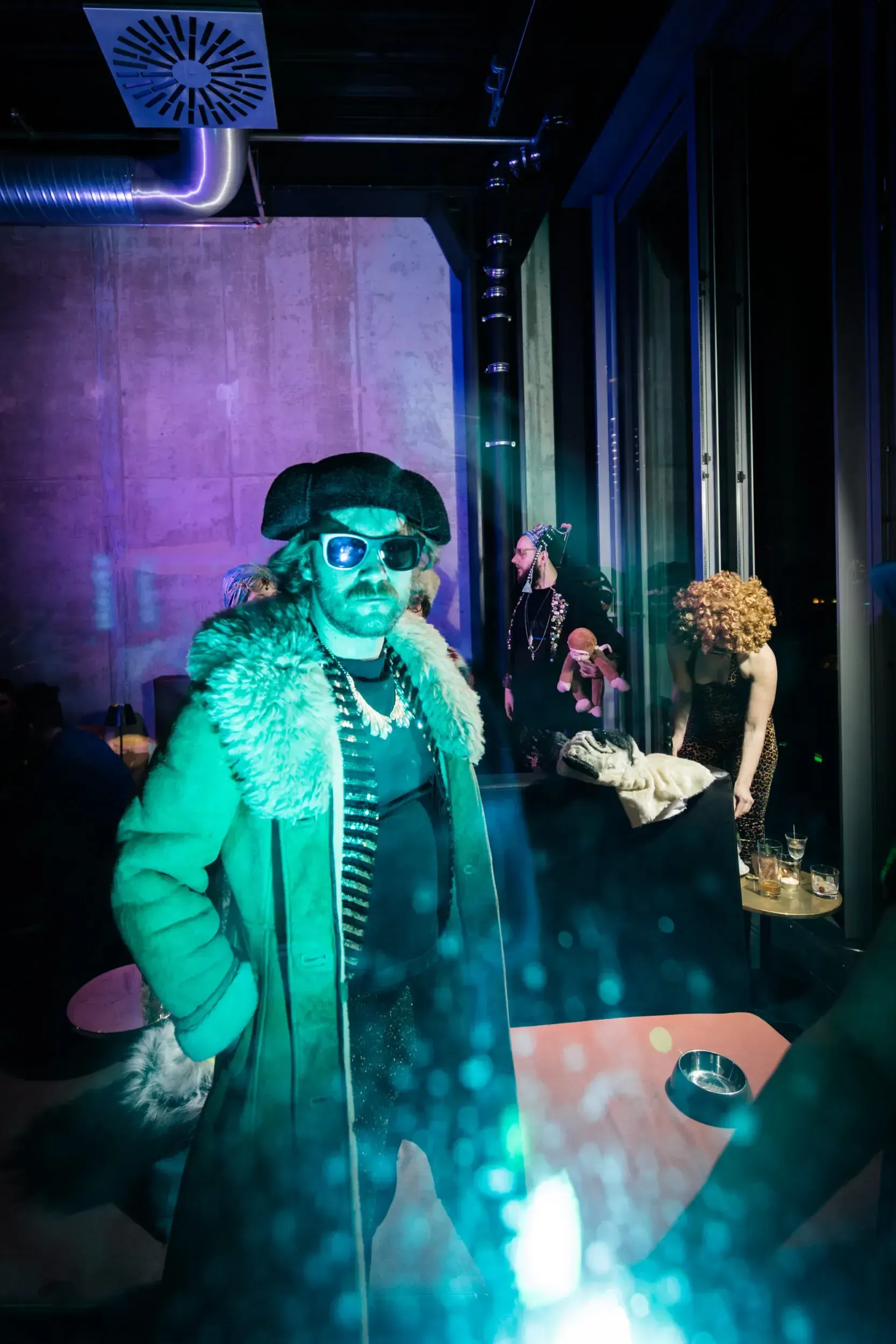 Guest in a fur-collared coat and sunglasses lit in green, other costumed guests in the background.