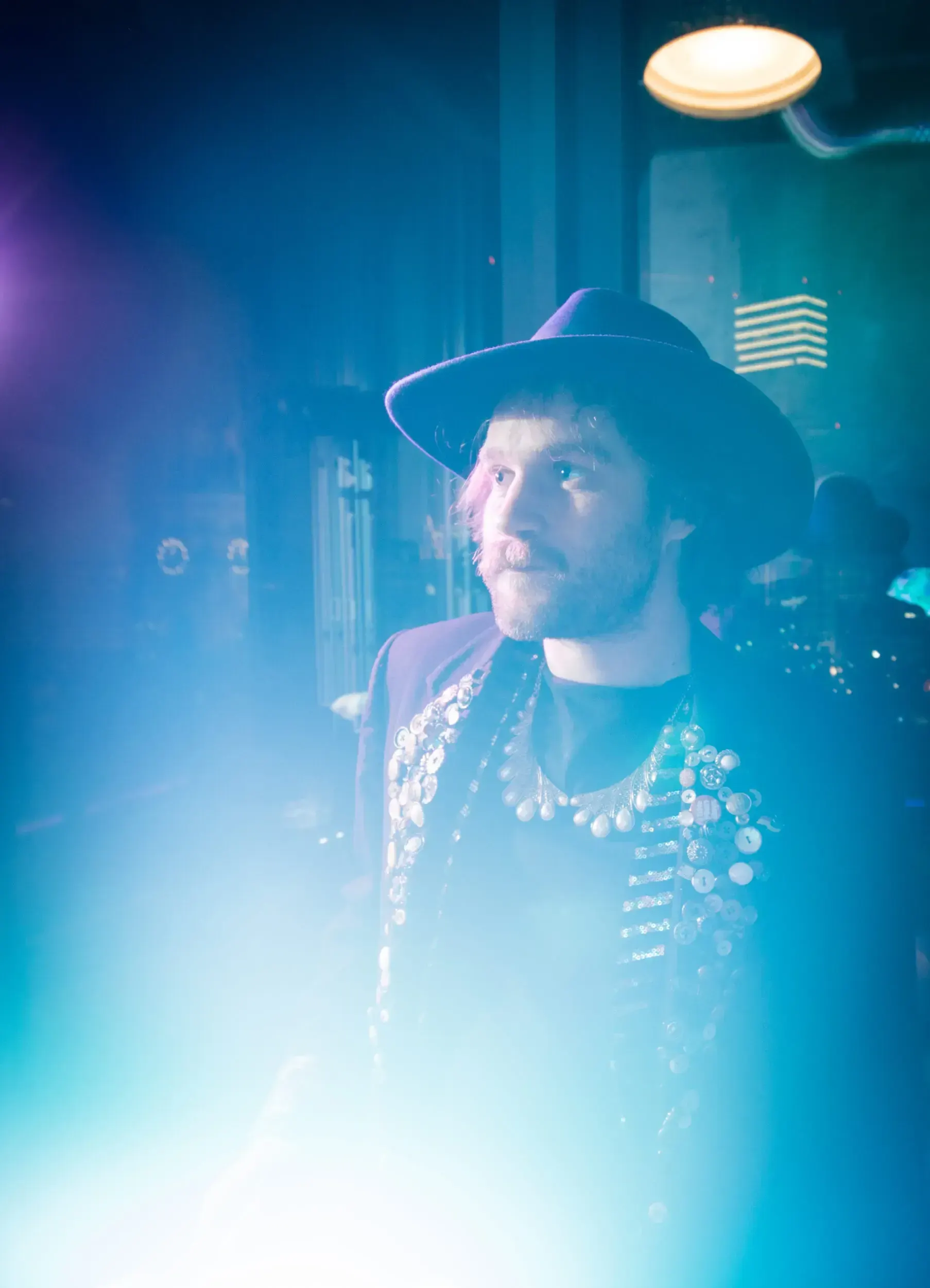 Man in a hat and layered necklaces lit in blue haze at the party.