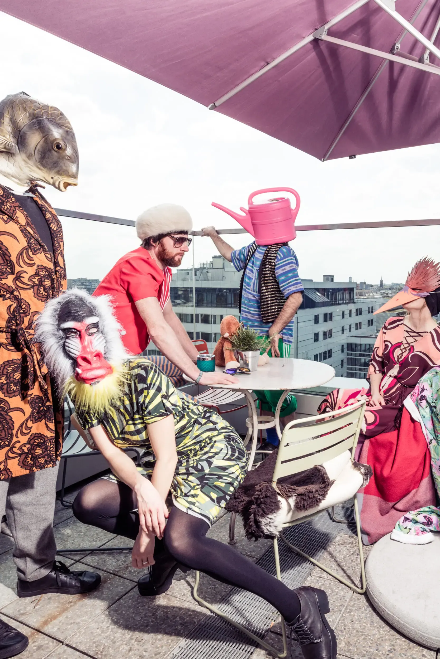 Guests in monkey masks lounging together on a rooftop terrace under a pink umbrella.