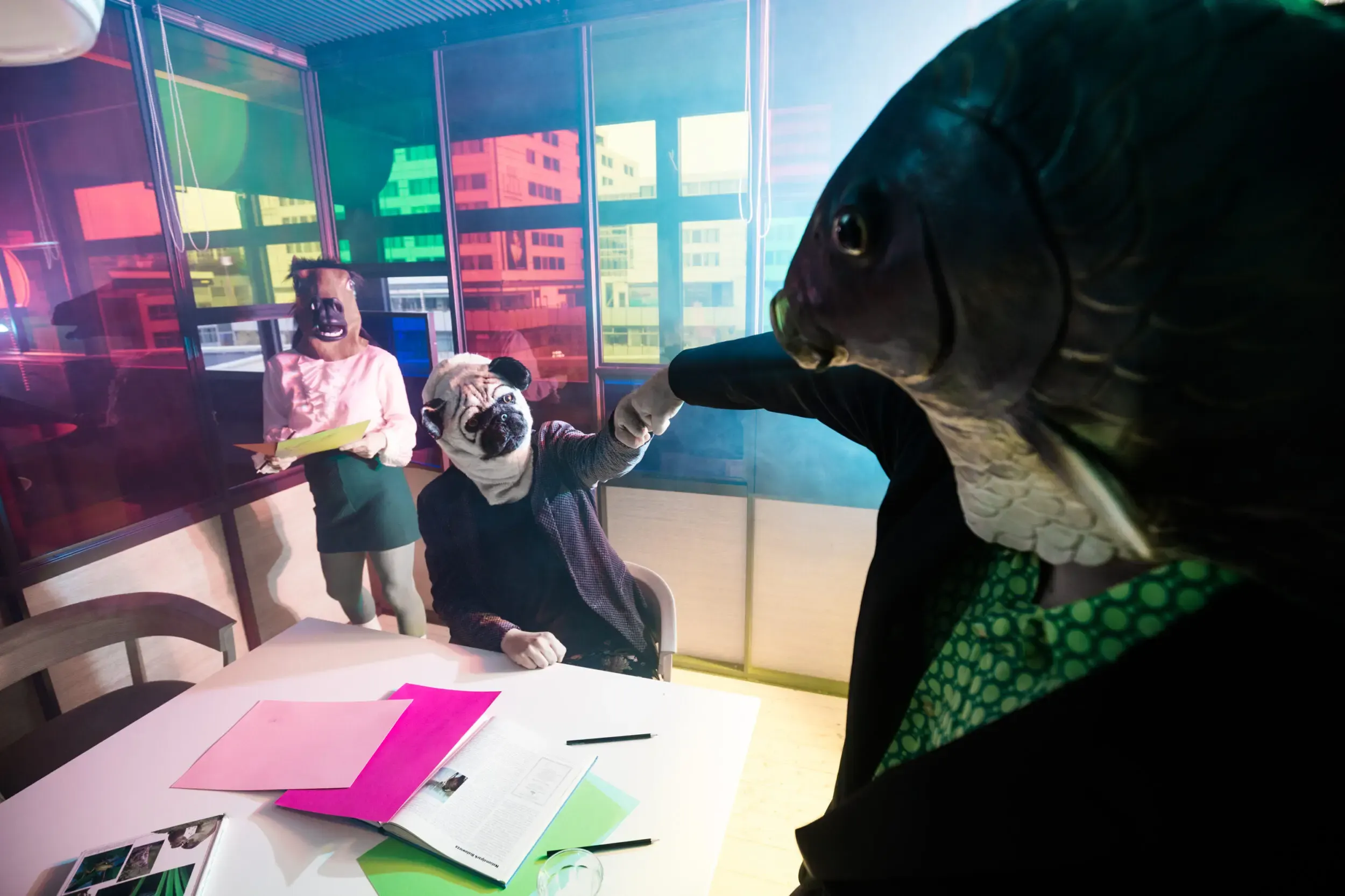 Costumed guests seated at an office desk under green lighting, one wearing a watering-can mask.