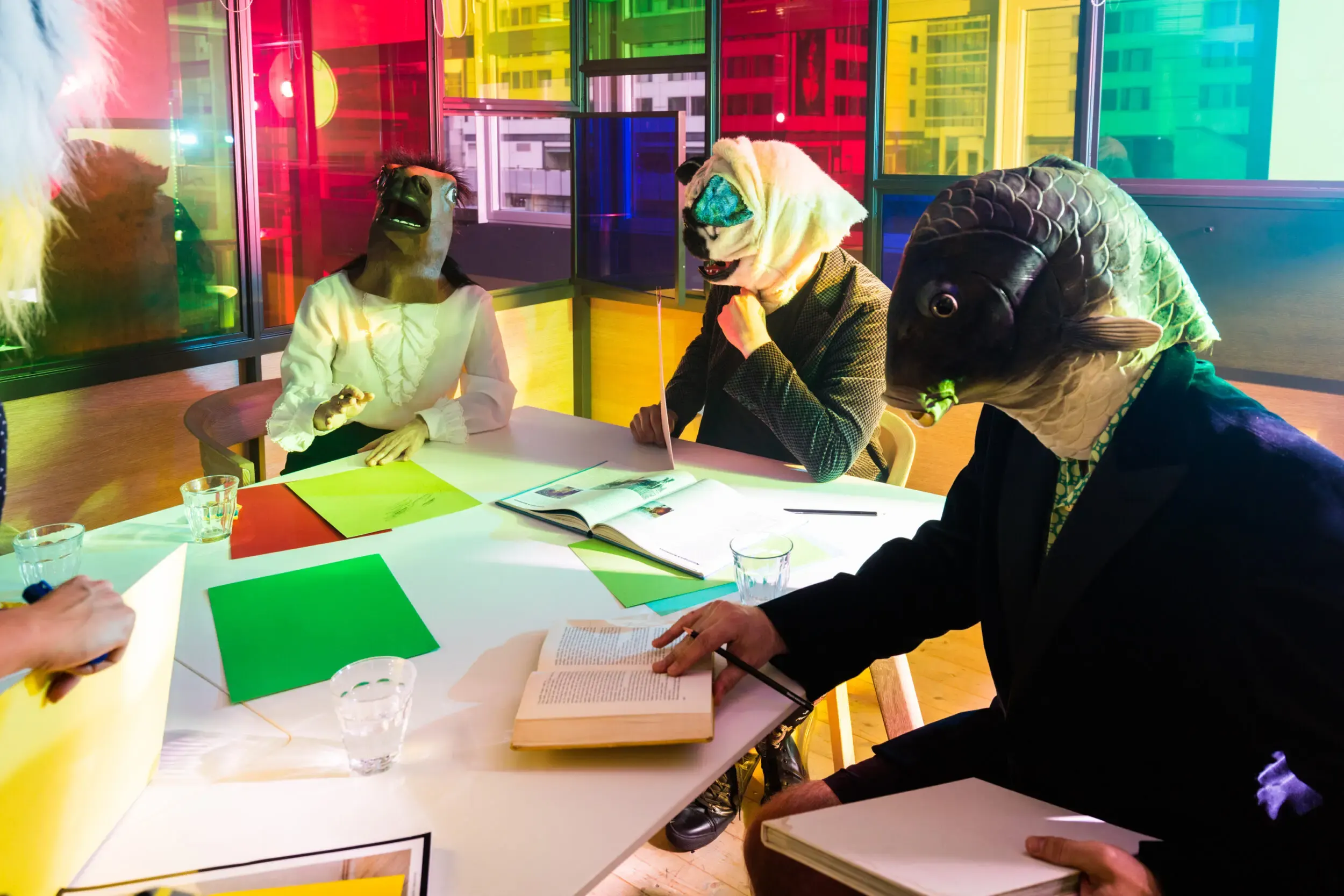 Guests in horse, sheep and fish masks seated around an office meeting table lit in rainbow colours.