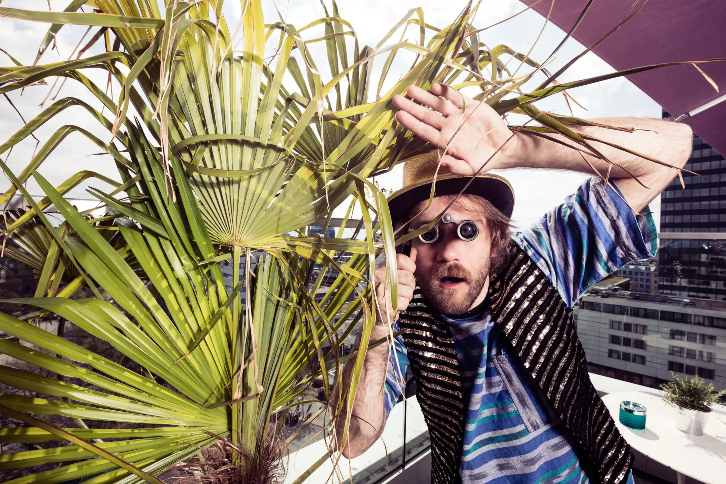 Guest in a hat and sunglasses posing among palm leaves on a rooftop, skyline behind.
