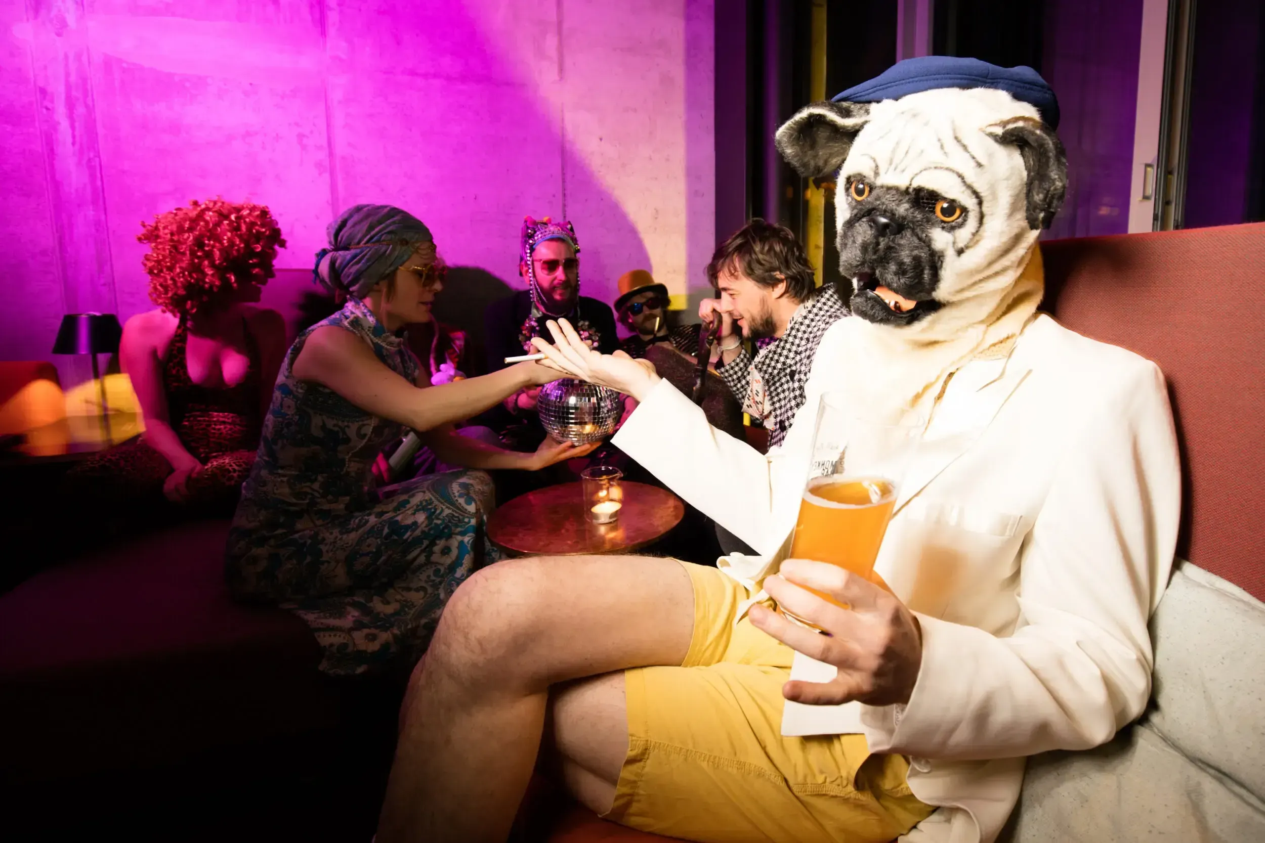 Person in a pug mask and white suit holding a drink, seated among other costumed party guests.