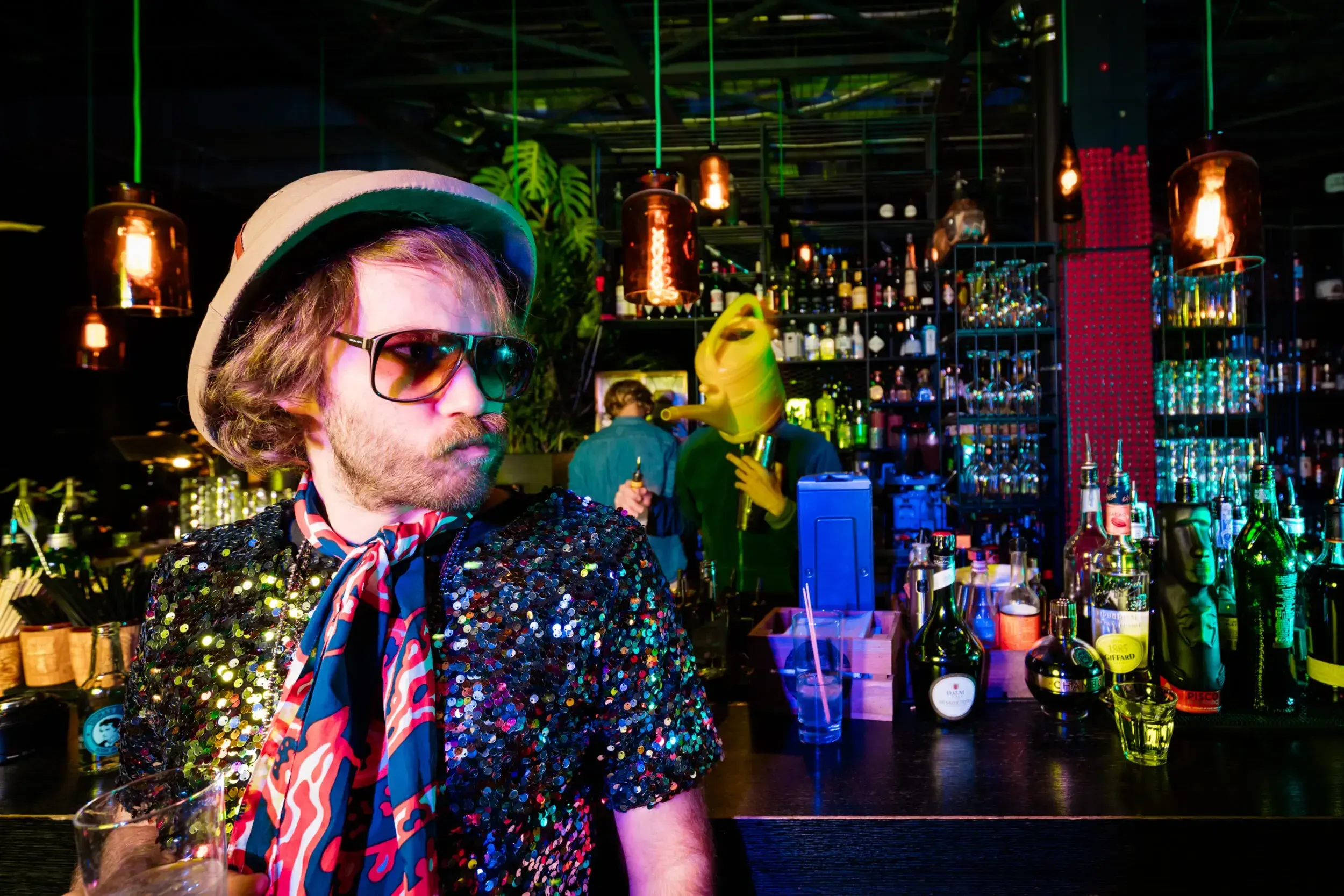 Man in a sequin jacket and headscarf at a bar, a guest in a watering-can mask smoking behind him.