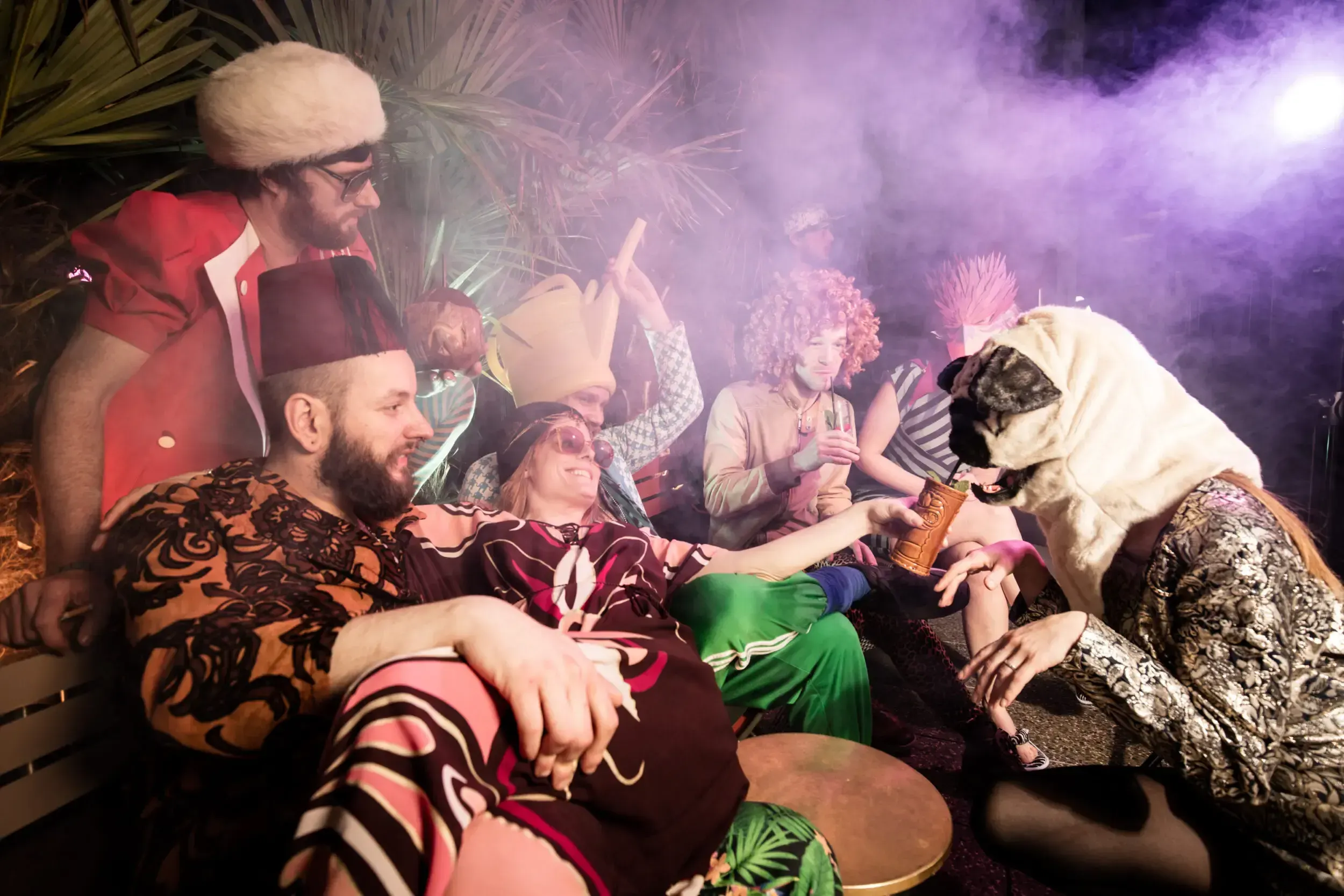 Costumed guests on the terrace passing a drink to a person in a pug mask amid haze.