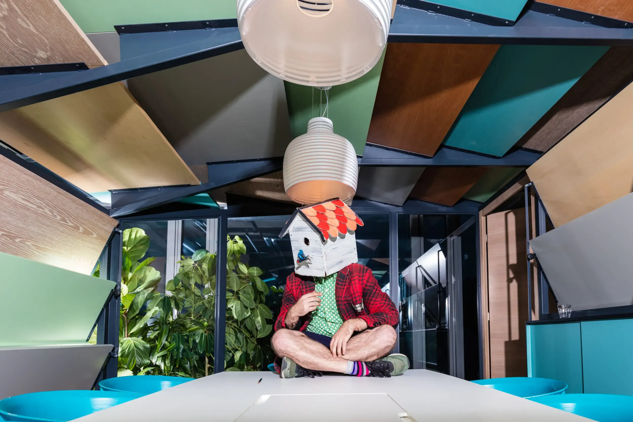 Guest in a watering-can mask upside down beneath paper lanterns in a colourful room.