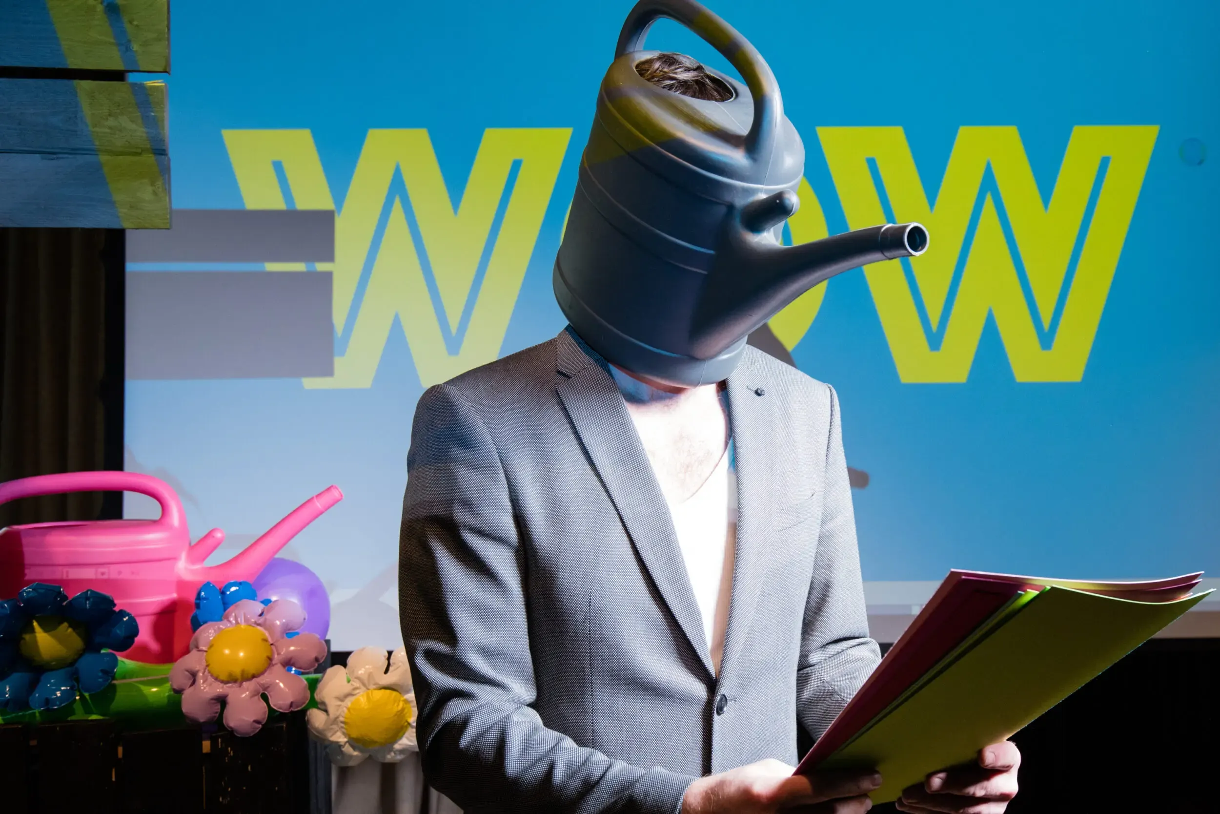 Guest in a watering-can mask and grey suit reading a folder in front of a 'WOW' backdrop.
