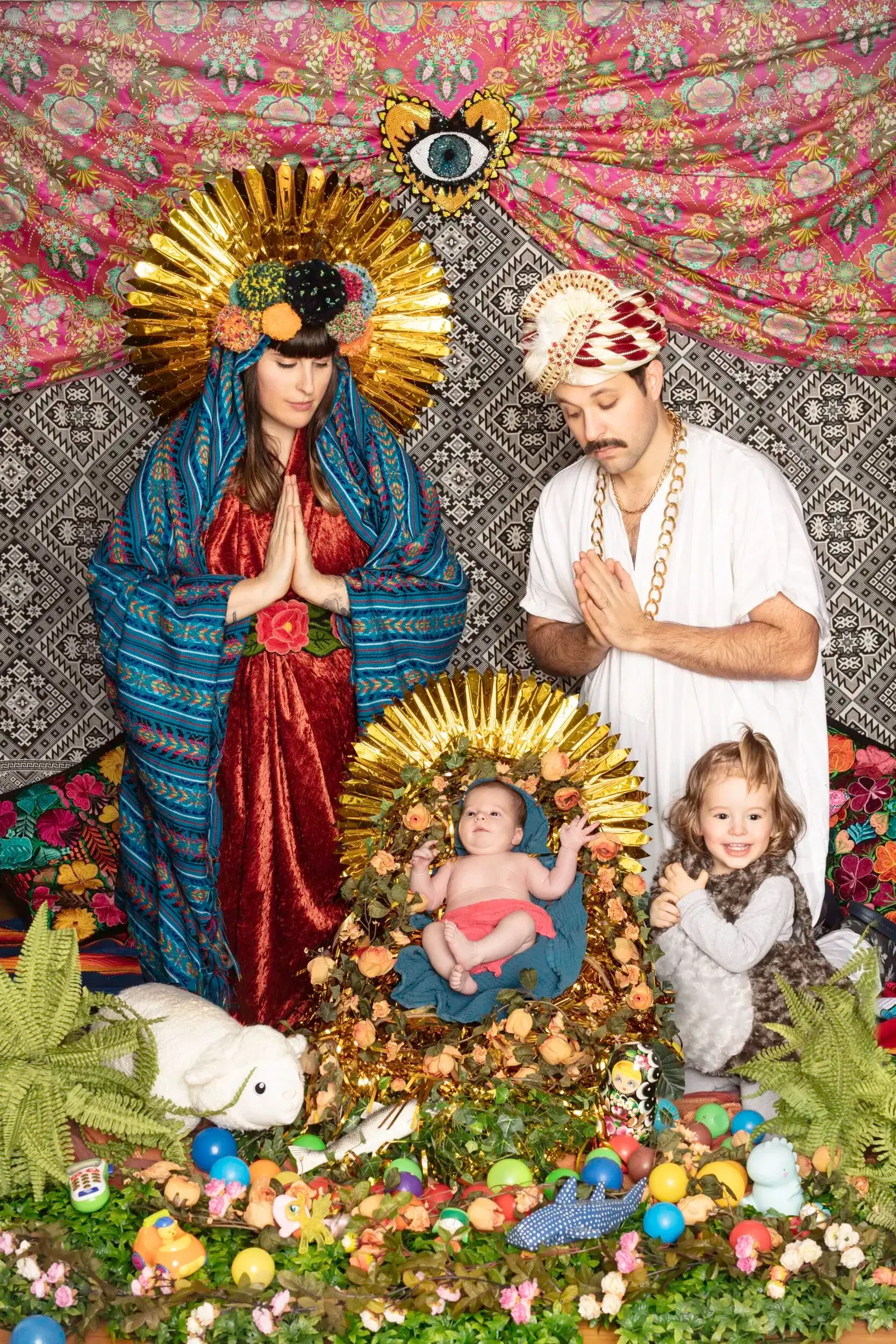 Family posed in a colorful nativity-style portrait with golden halos, silk robes and toys.