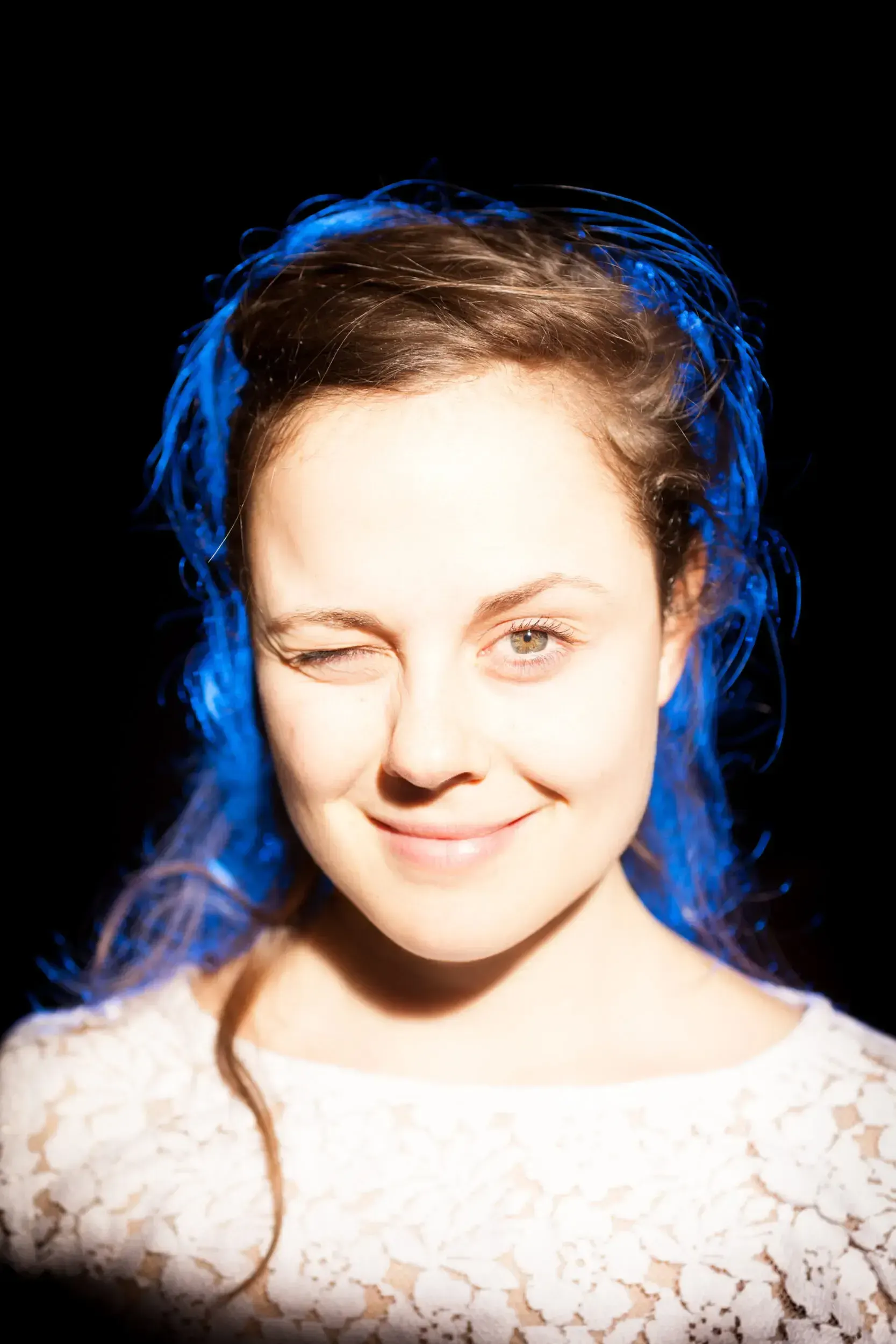 Winking portrait of Elisa Plüss smiling in sunlight, her hair lit blue against a black background.