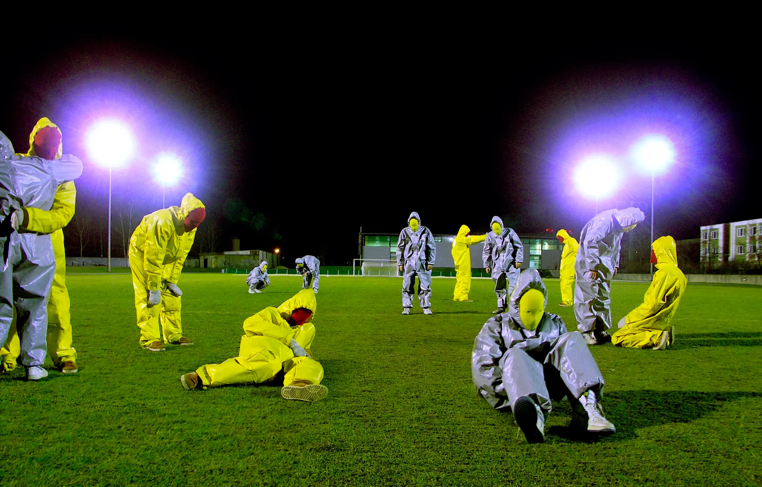 Gruppe im Schutzanzug auf einem nächtlich beleuchteten Sportfeld, graugelb™
