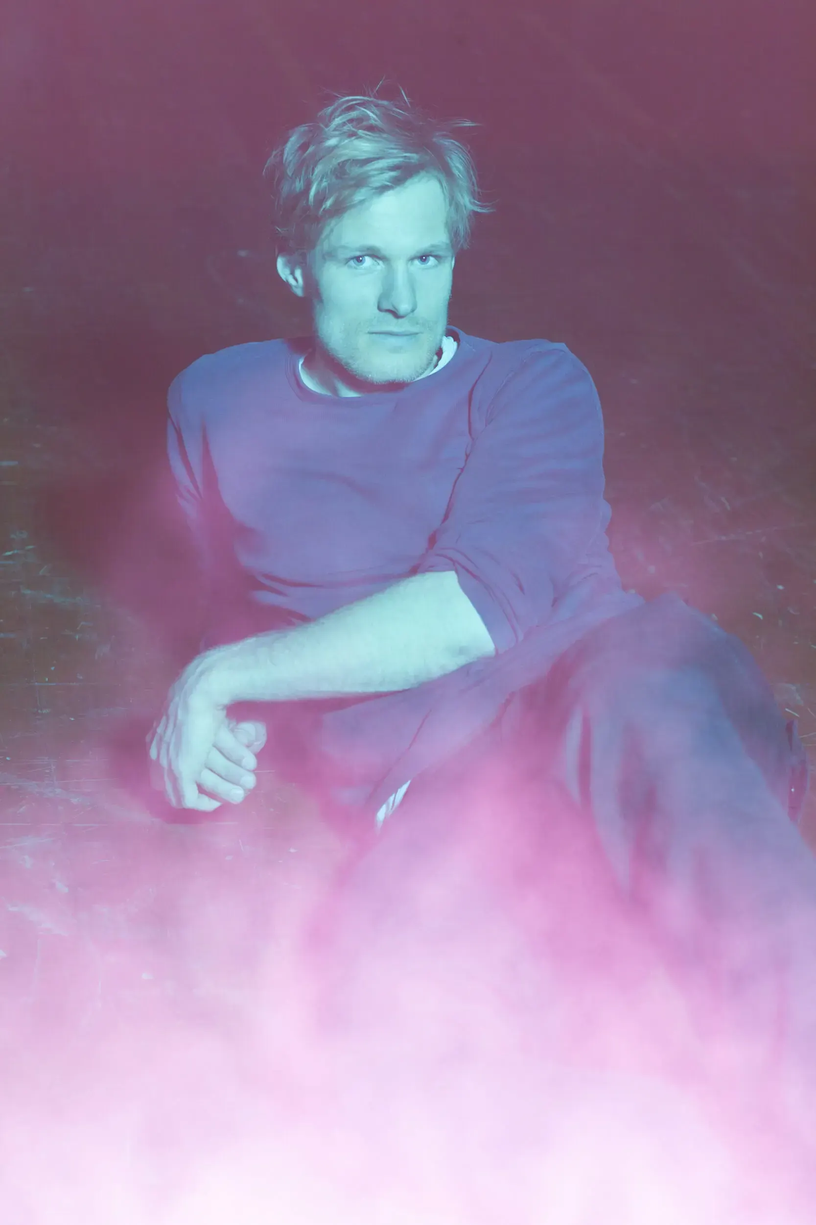 Portrait of actor Jirka Zett sitting on the floor, lit in blue and pink haze.