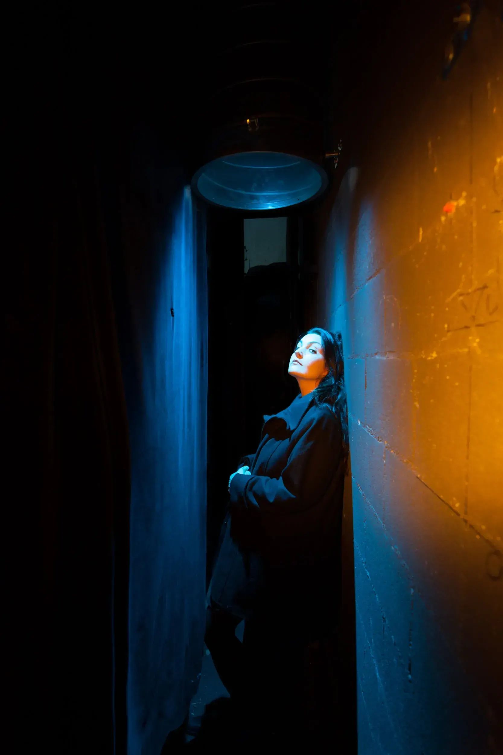 Portrait of actress Julia Kreusch leaning against a wall in a narrow alley lit in blue and orange.