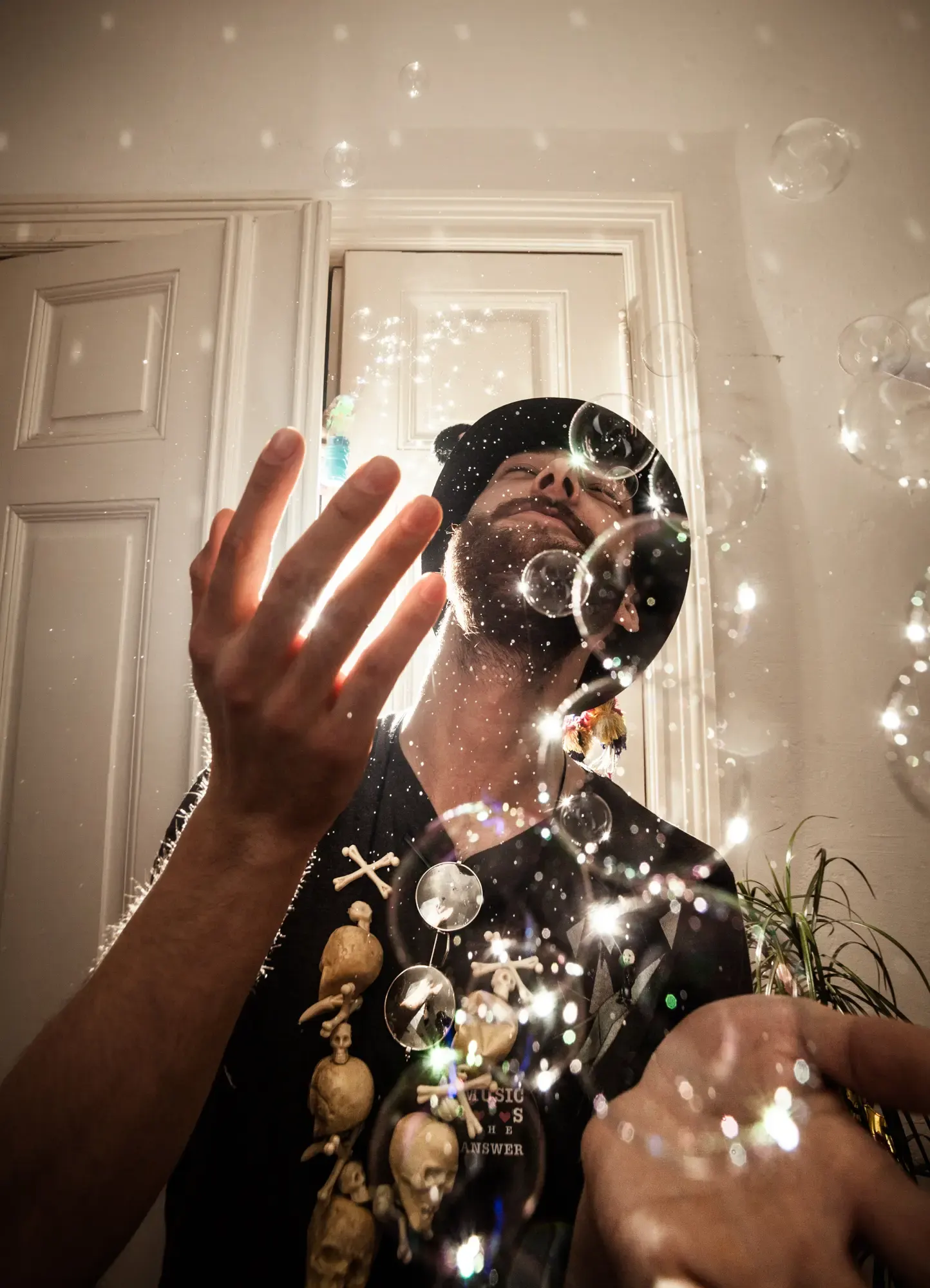 A person with a black hat and shirt, surrounded by shimmering soap bubbles in a bright indoor setting.