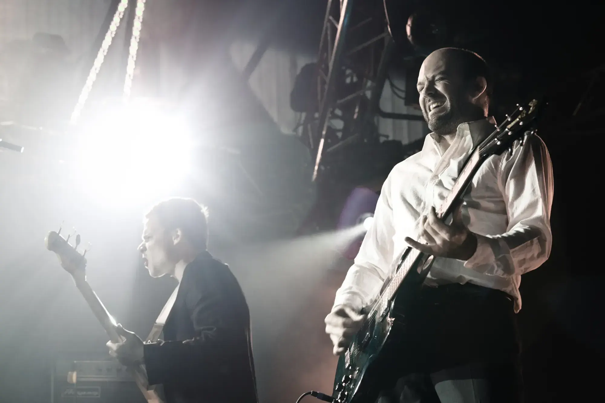 Bassist and guitarist performing live on stage, one singing into a spotlight.