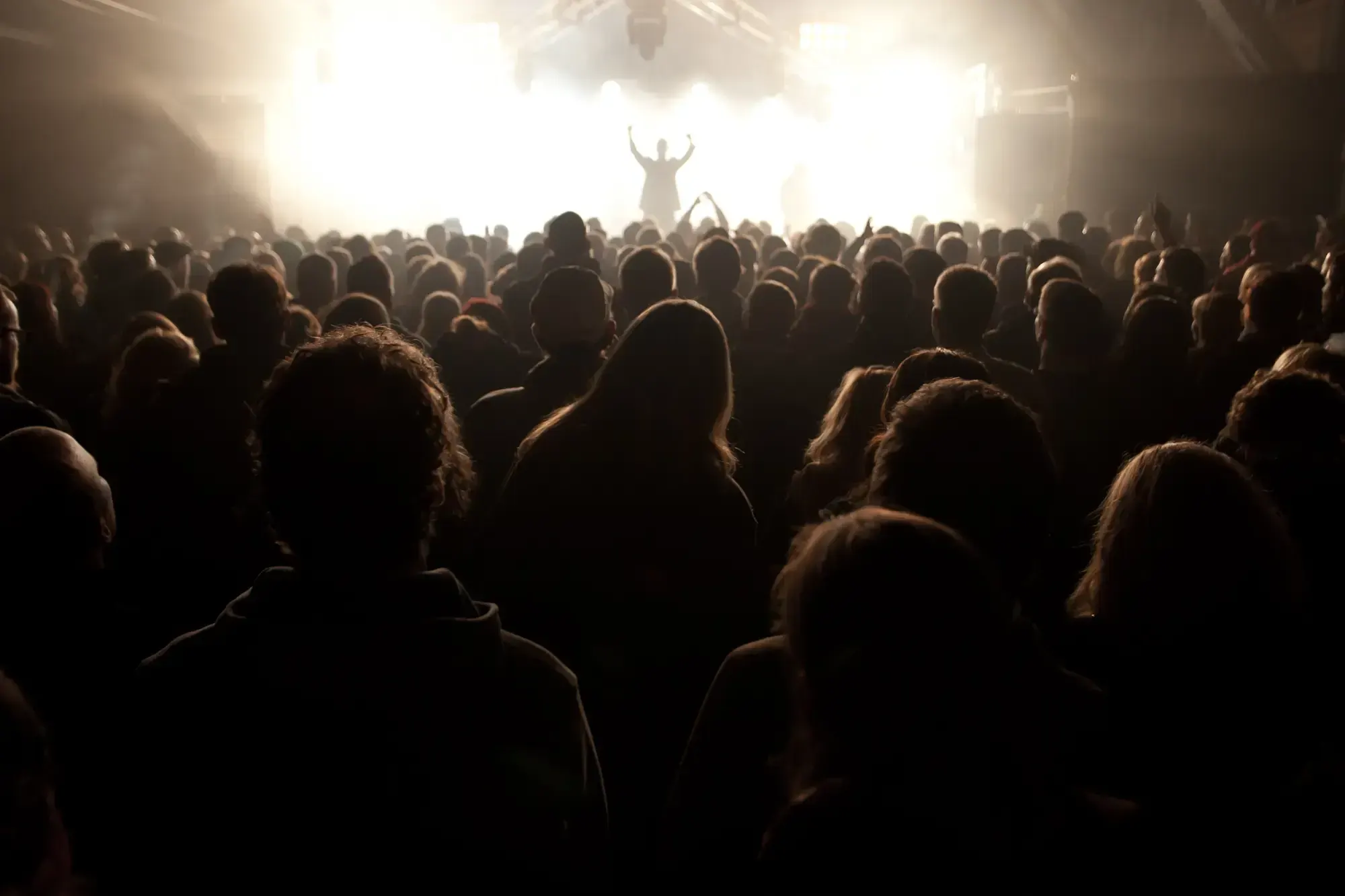 View from the stage over a crowd watching a concert, arms raised silhouetted against bright lights.