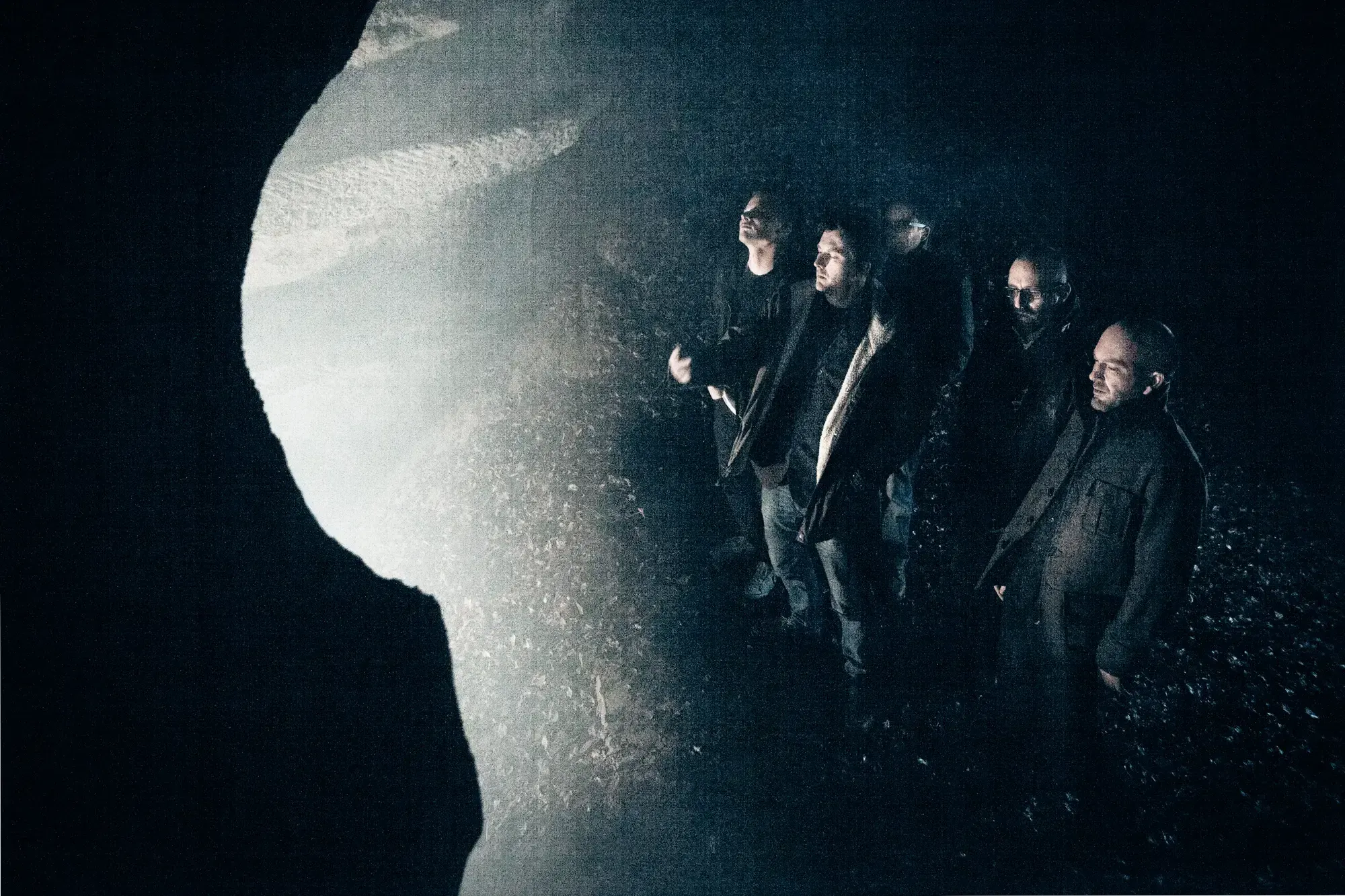 Band standing in profile inside a dark cave-like space, lit from one side.