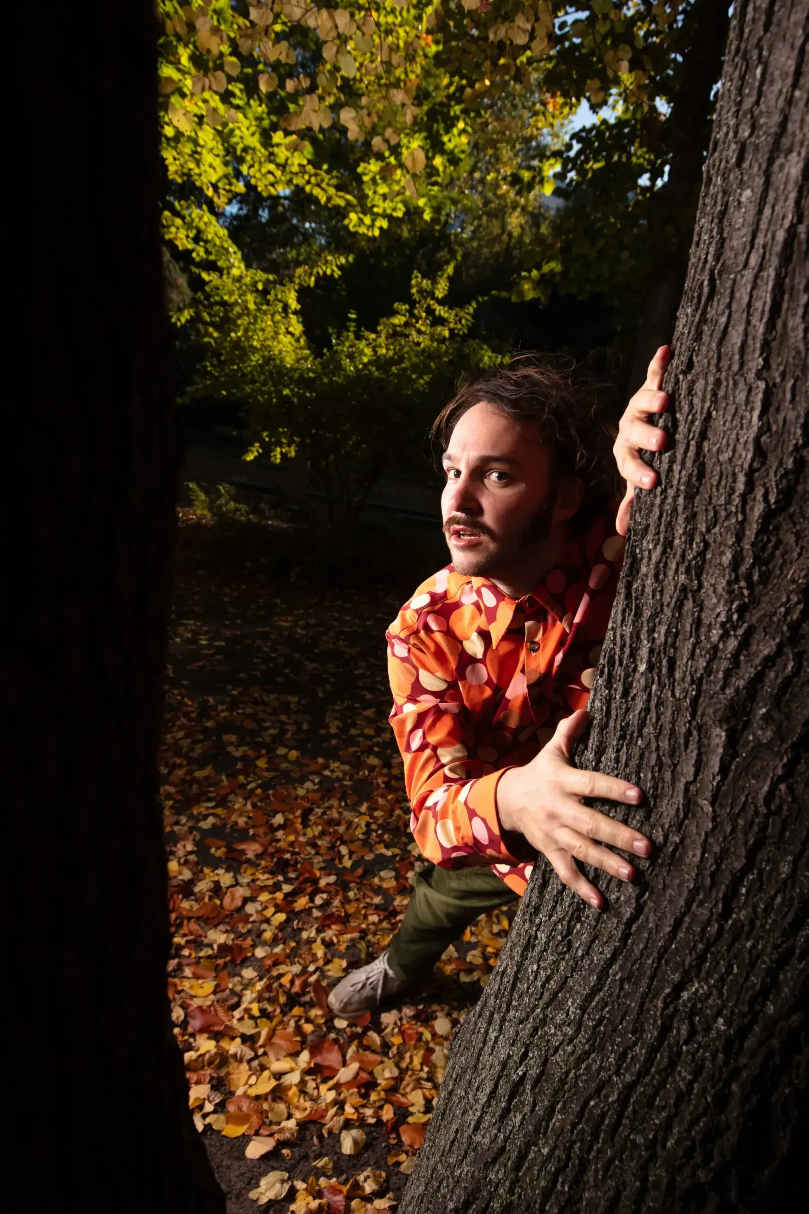 Man in a retro orange patterned shirt peering out from behind a tree trunk in an autumn park.