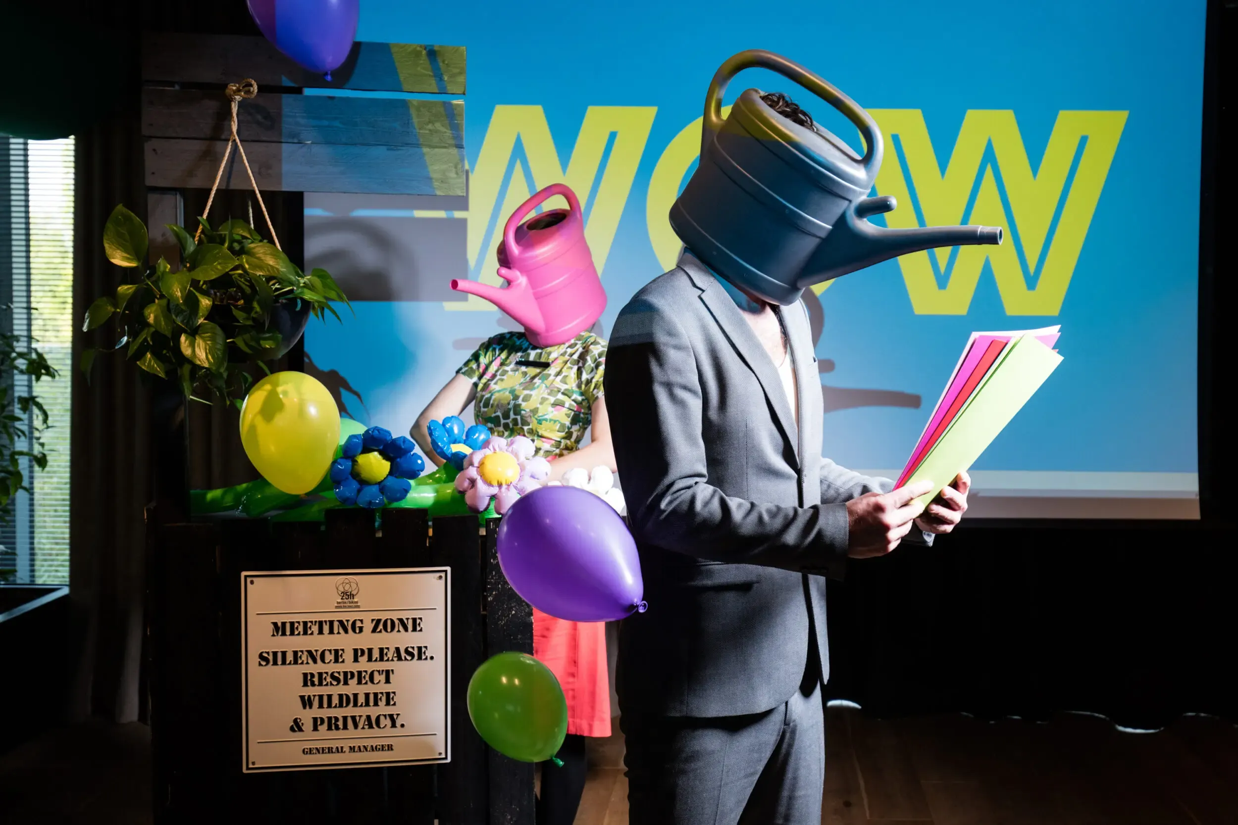 Gast mit Giesskannen-Maske und grauem Anzug vor einem „Meeting Zone“-Schild