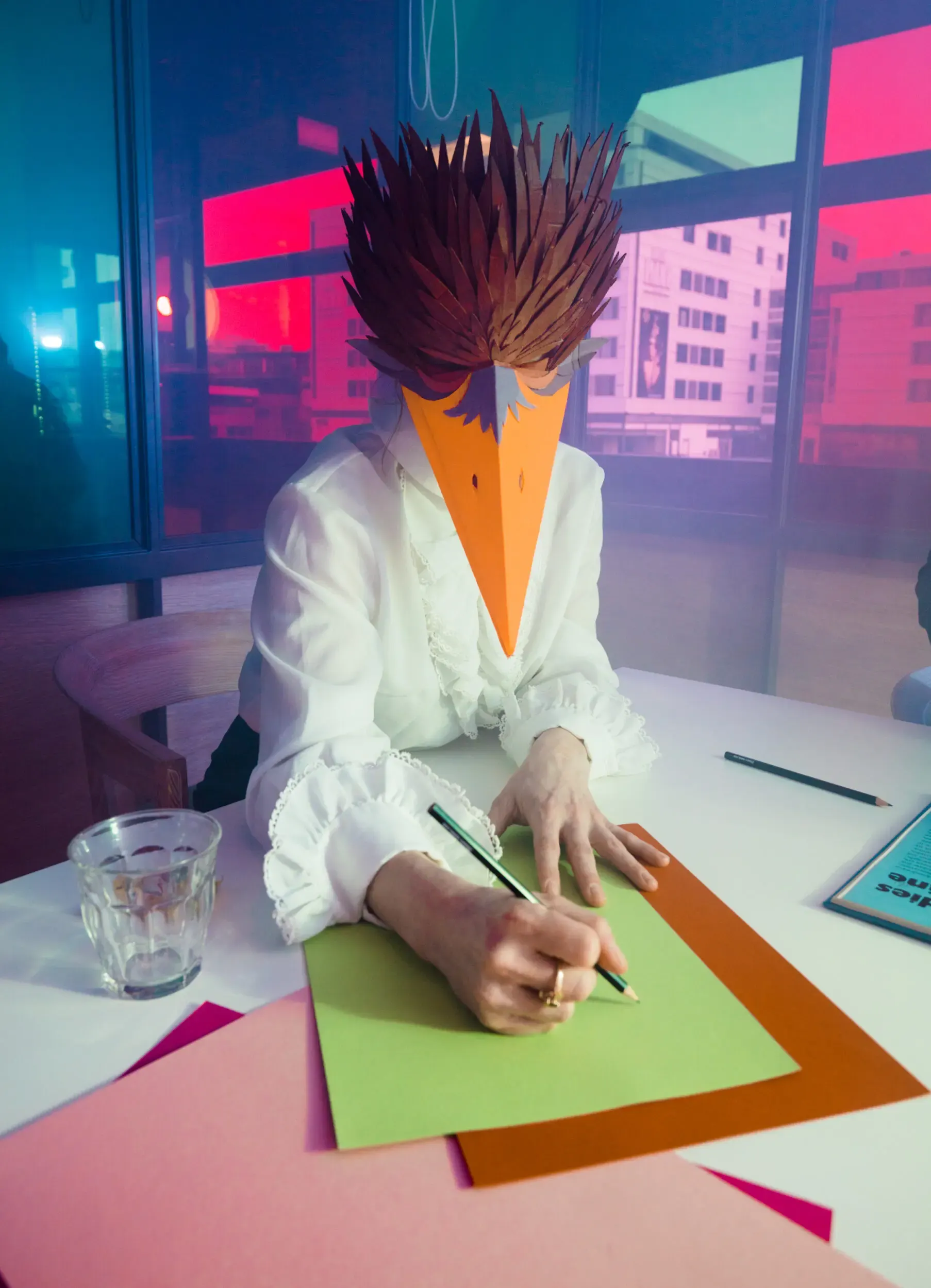 Person mit vogelartiger Maske sitzt schreibend an einem Schreibtisch, neonbeleuchtetes Büro bei Nacht