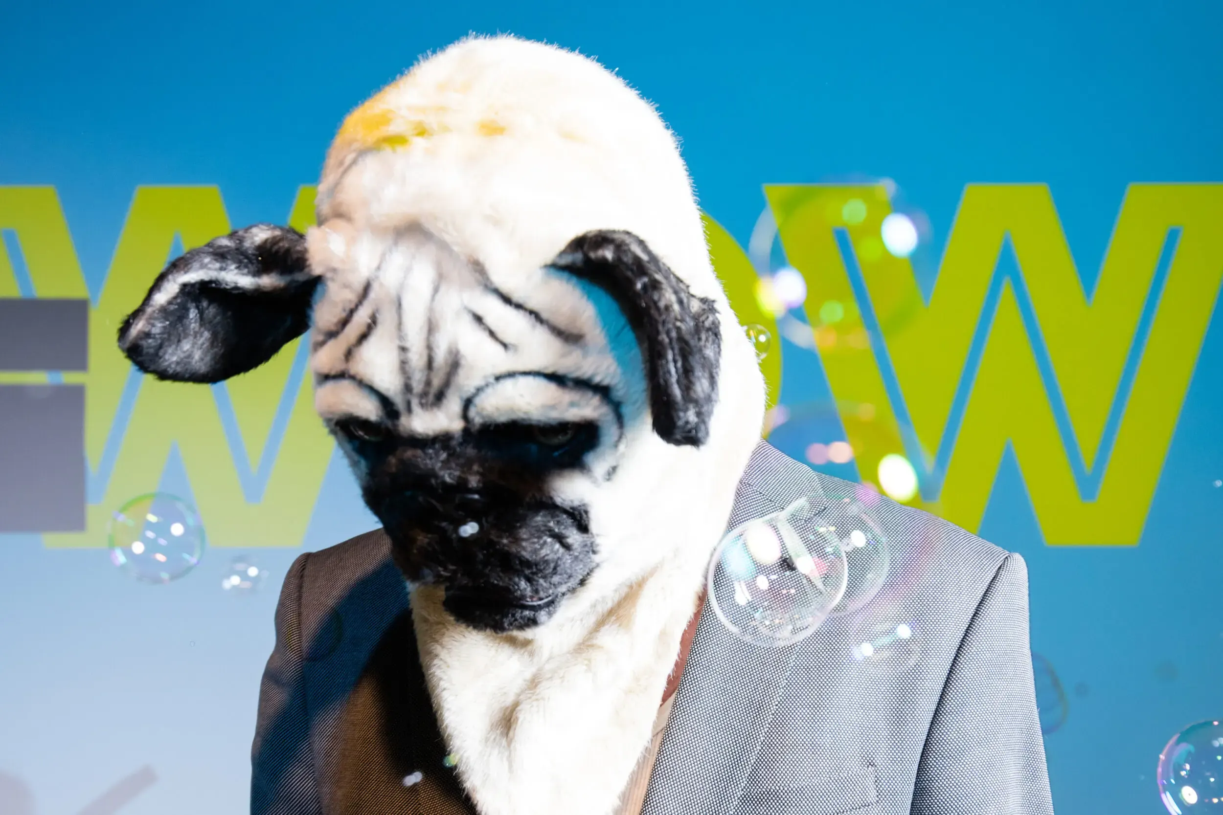 Gast mit Mops-Maske und grauem Anzug bläst Seifenblasen vor „WOW“-Kulisse