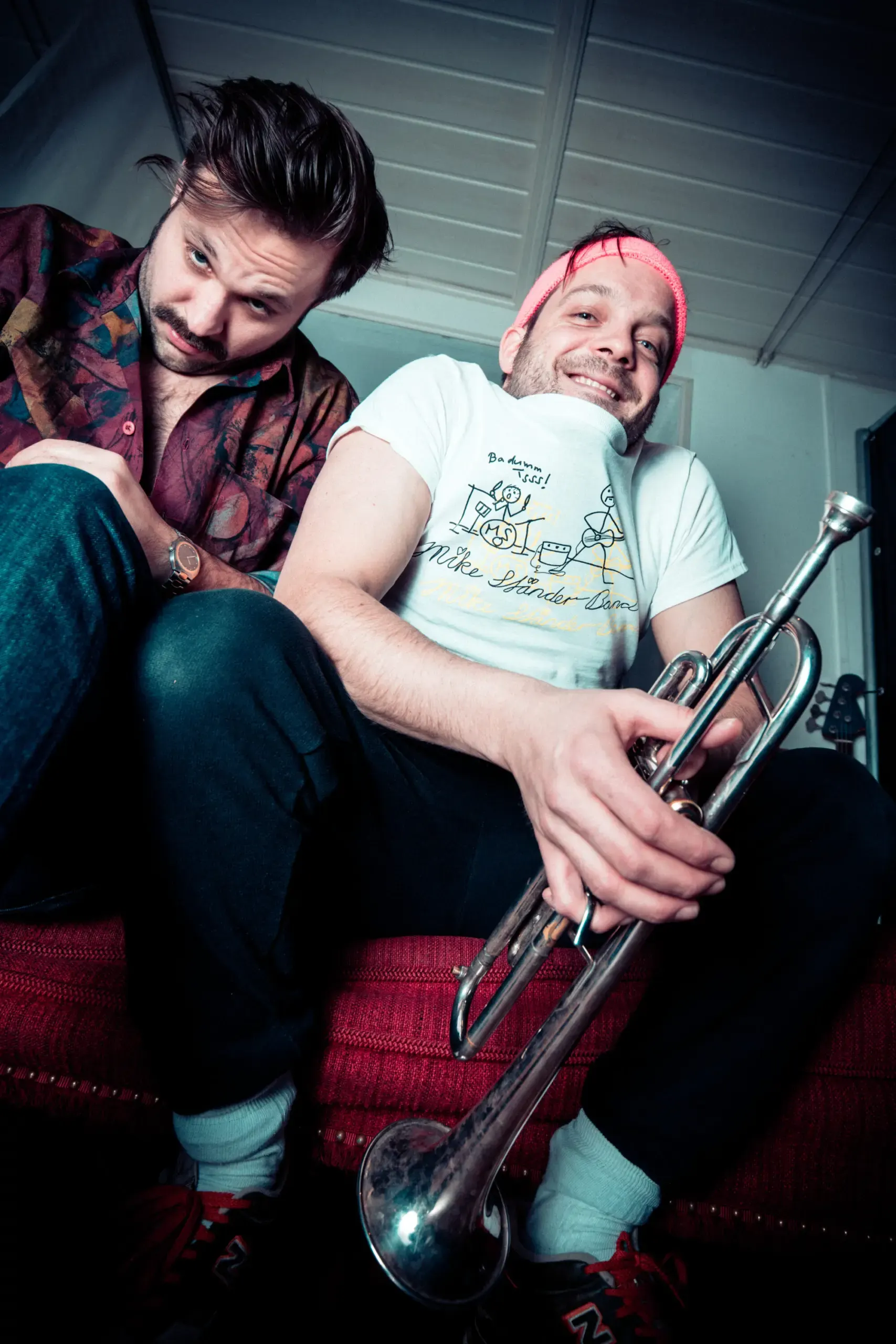 Bandduo sitzt lächelnd mit Trompete auf rotem Sofa