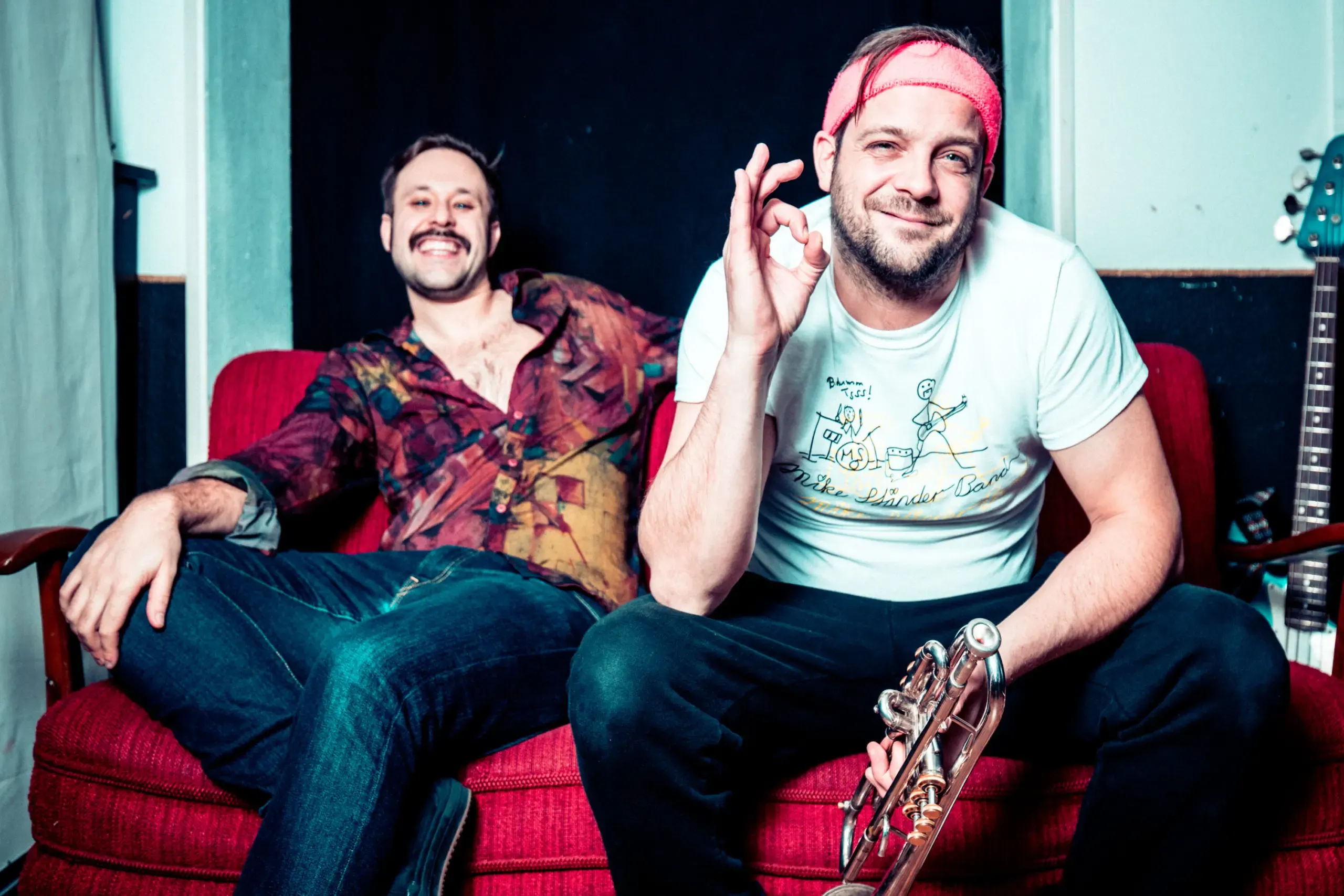 Bandduo posiert auf rotem Sofa, einer zeigt OK-Zeichen