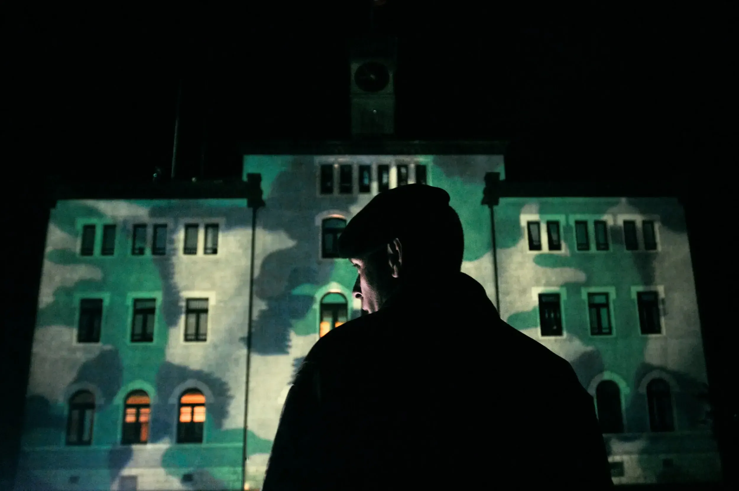 Silhouette of a soldier in a cap standing before a barracks building projected with a camouflage pattern at night.
