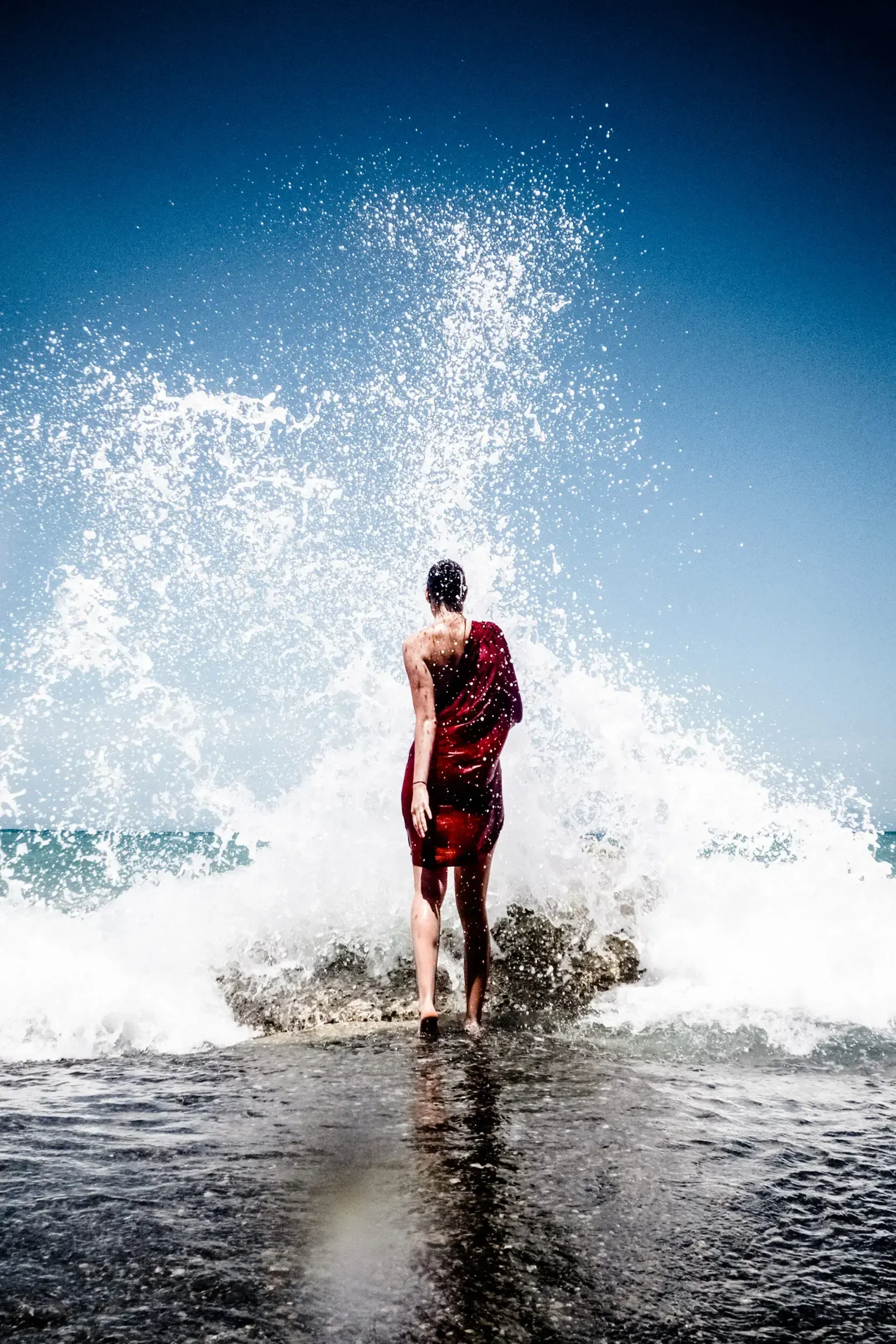 Woman in a red dress standing in the surf as a wave crashes behind her.
