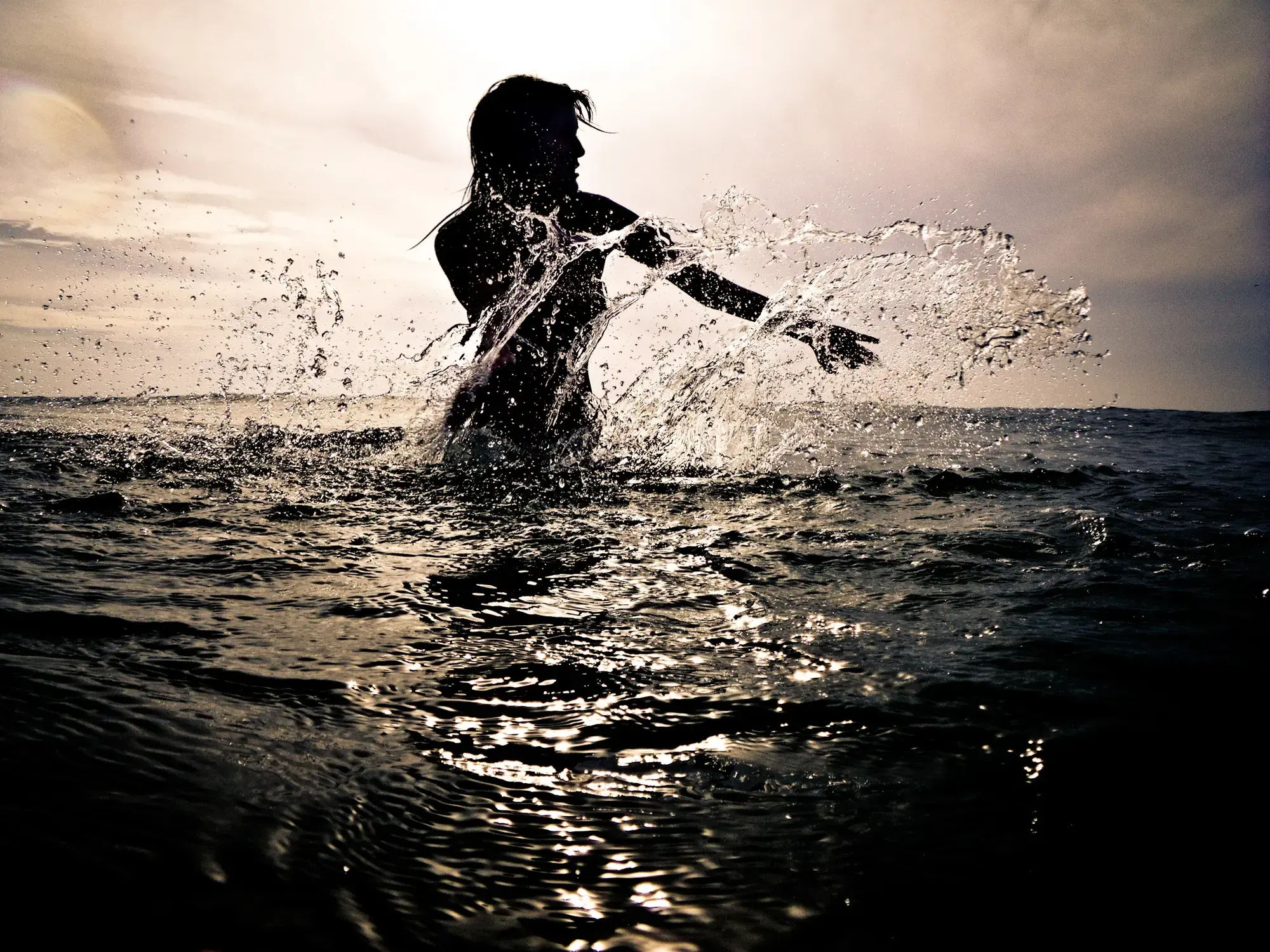 Silhouette of a woman splashing water with her arm while standing in the sea at sunset.