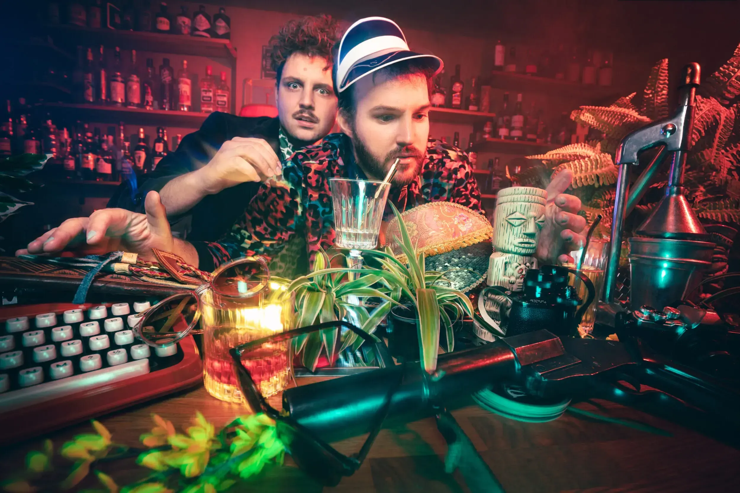 Two men behind a tiki bar counter cluttered with cocktails, plants and vintage props.