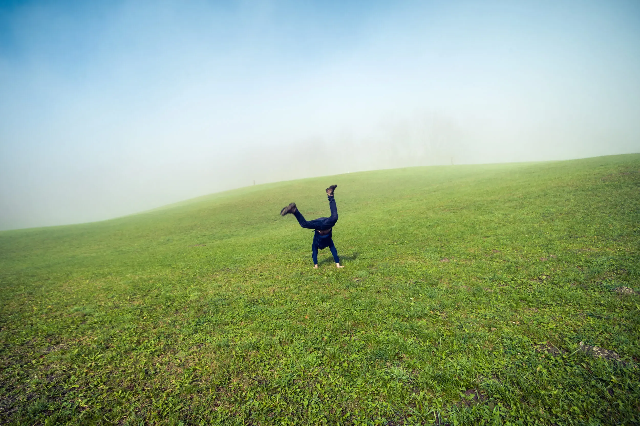 A person doing a handstand on lush green grass under a foggy sky.