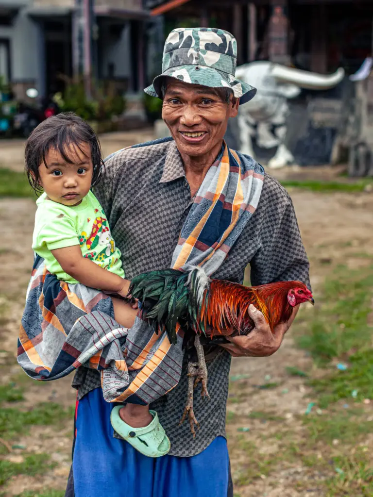 A man wearing a camouflage hat holds a rooster in one hand while carrying a child in the other.