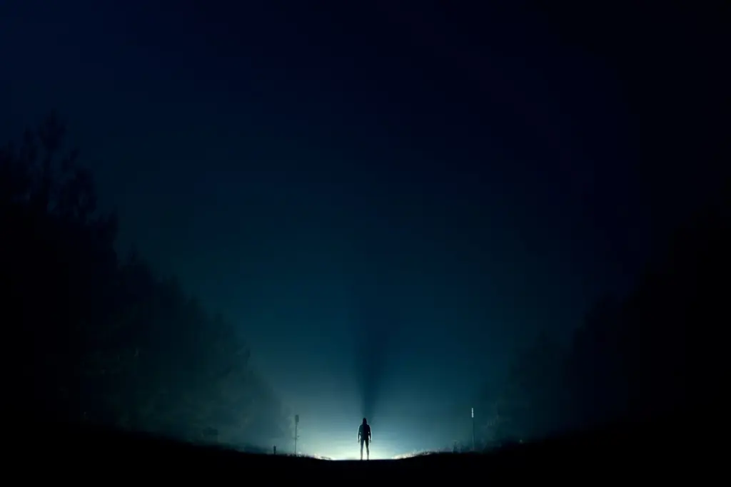 Silhouette of a person standing on a dark road, surrounded by fog and illuminated by a bright light in the background.