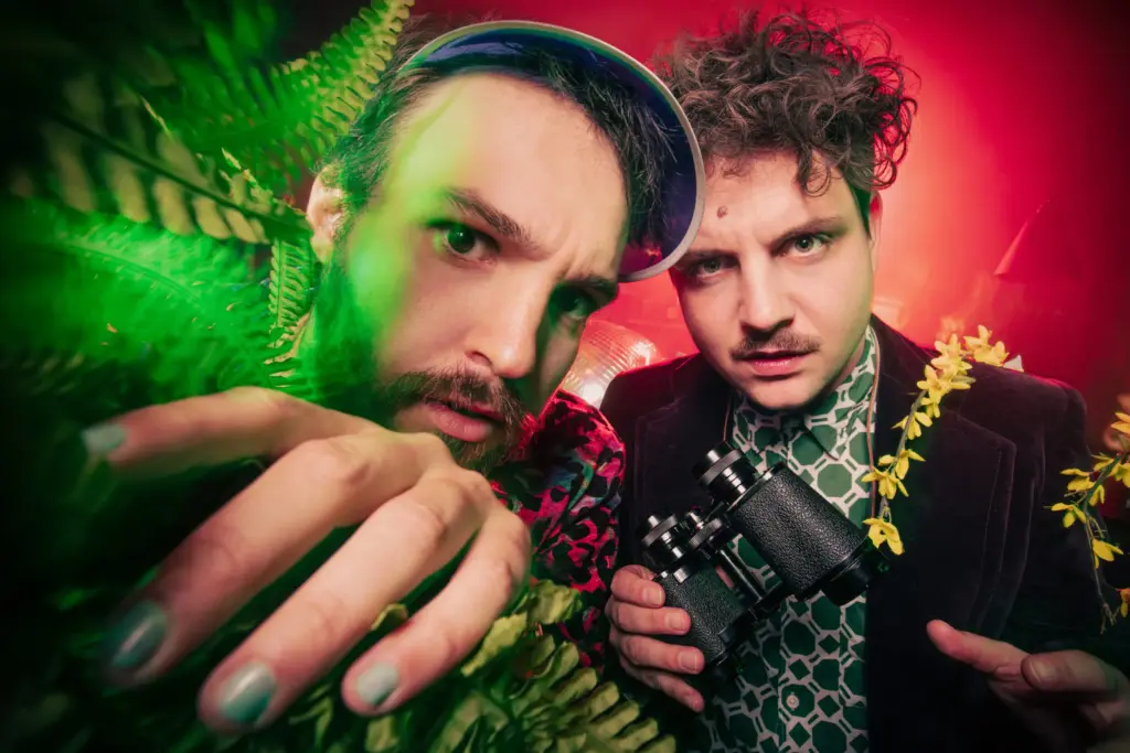 Two men surrounded by green ferns, one holding binoculars, wearing colorful outfits against a red background.