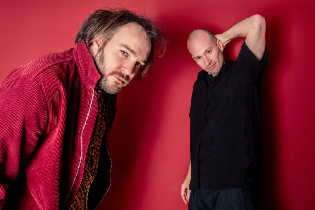 Two men posing against a red background, one in a red corduroy jacket and the other in a black shirt.