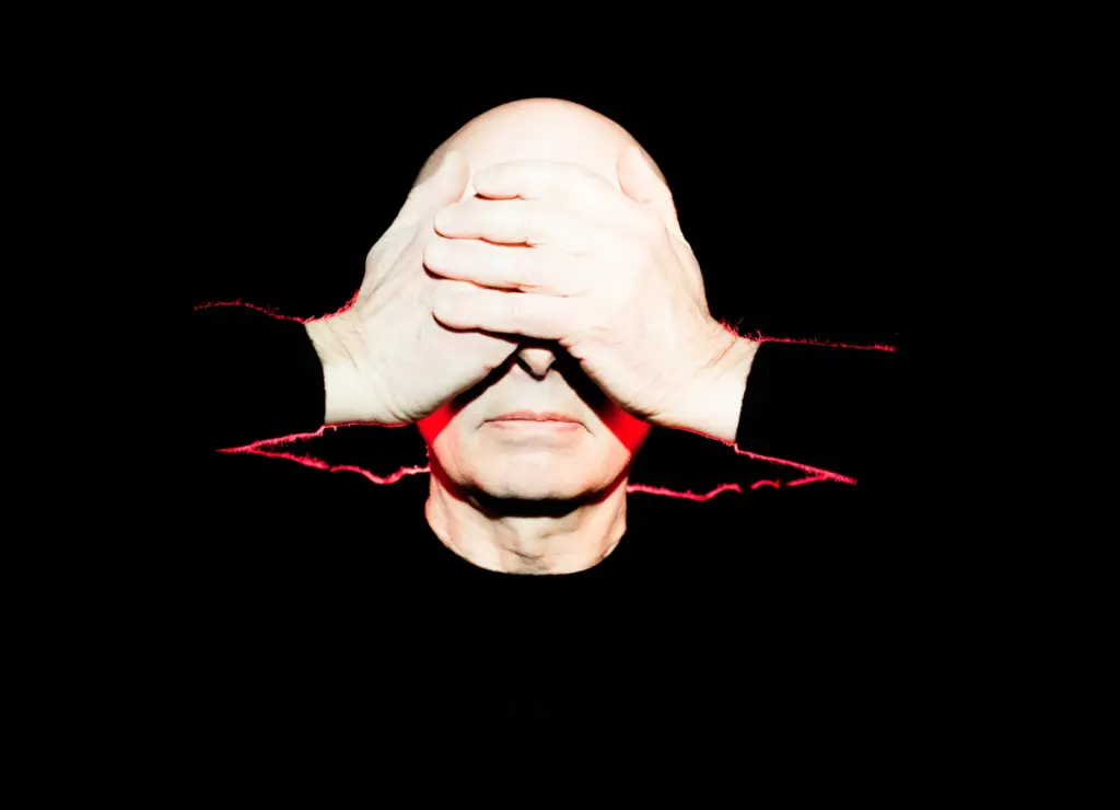 A person with a bald head covers their eyes with hands, wearing a black shirt, against a dark background.