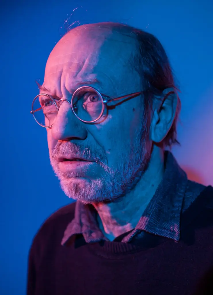 A man with glasses and a beard, wearing a dark sweater, illuminated by blue and purple lighting.