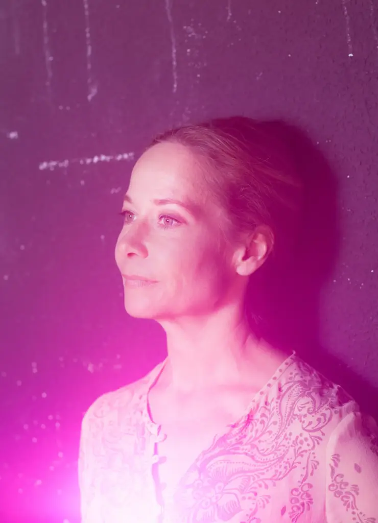 A woman in a light pink patterned blouse stands against a dark wall, illuminated by soft pink light.