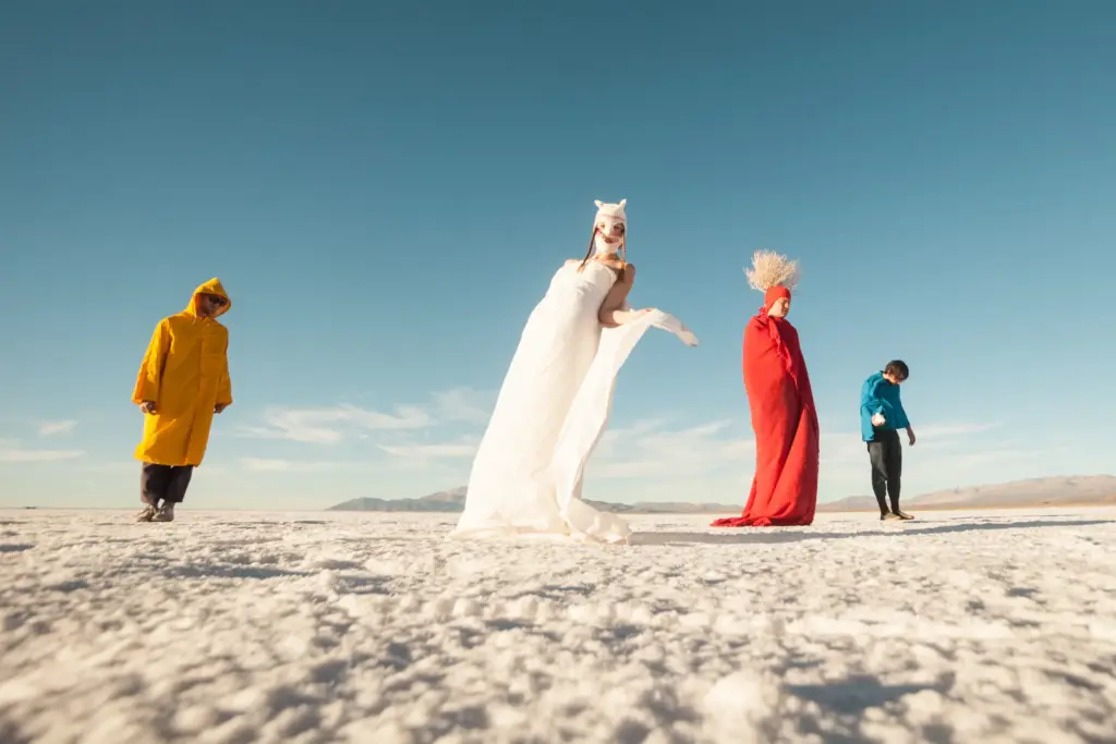 Four figures in colorful outfits on a white salt flat under a clear blue sky, showcasing unique styles and textures.