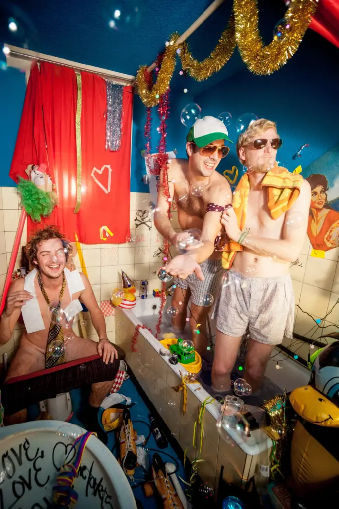 Three men in a colorful bathroom with bubbles, wearing shorts, surrounded by festive decorations and toys.