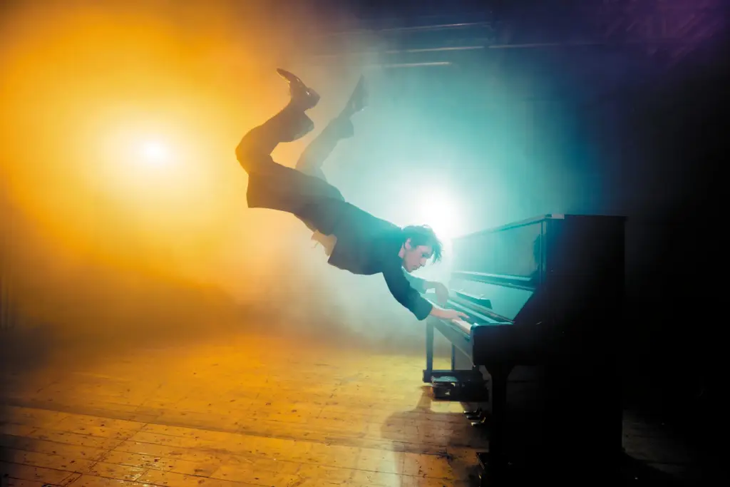 A man in a suit dramatically plays a black piano, surrounded by colorful fog and wooden floor.
