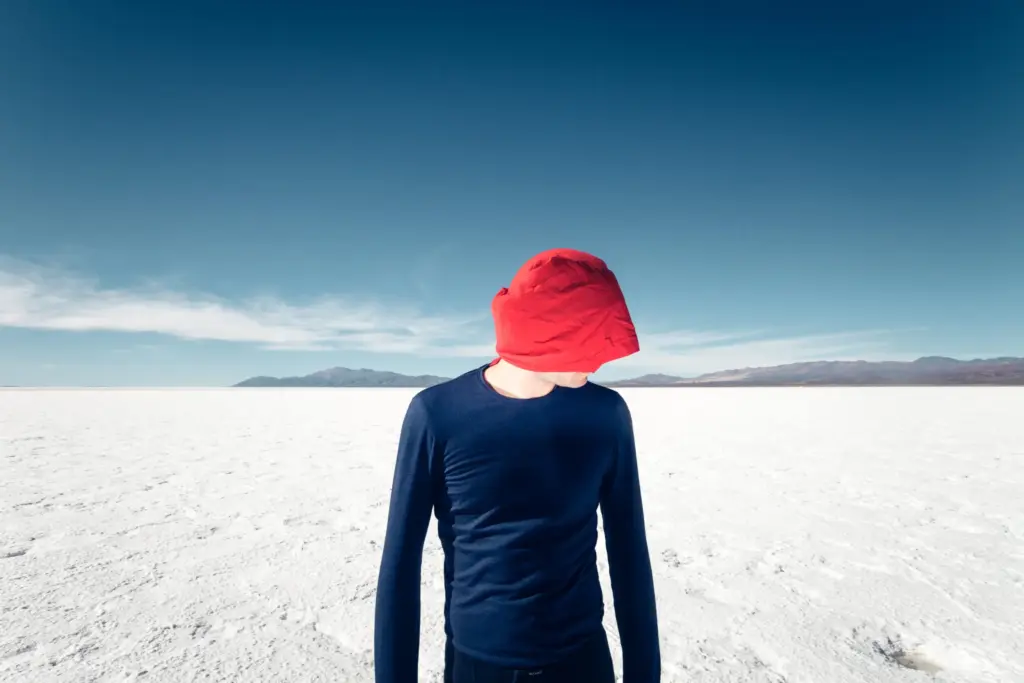 A person wearing a red fabric hood and a blue long-sleeve shirt stands on a white salt flat under a blue sky.