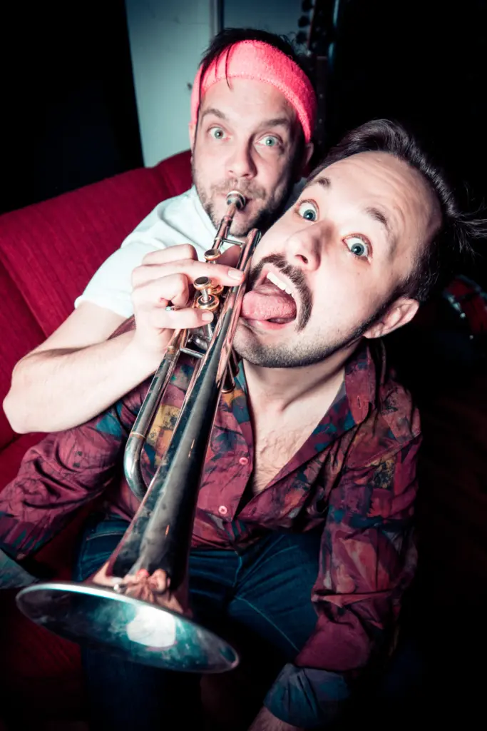 Two men, one playing a trumpet and the other making a playful face, both wearing colourful clothing.