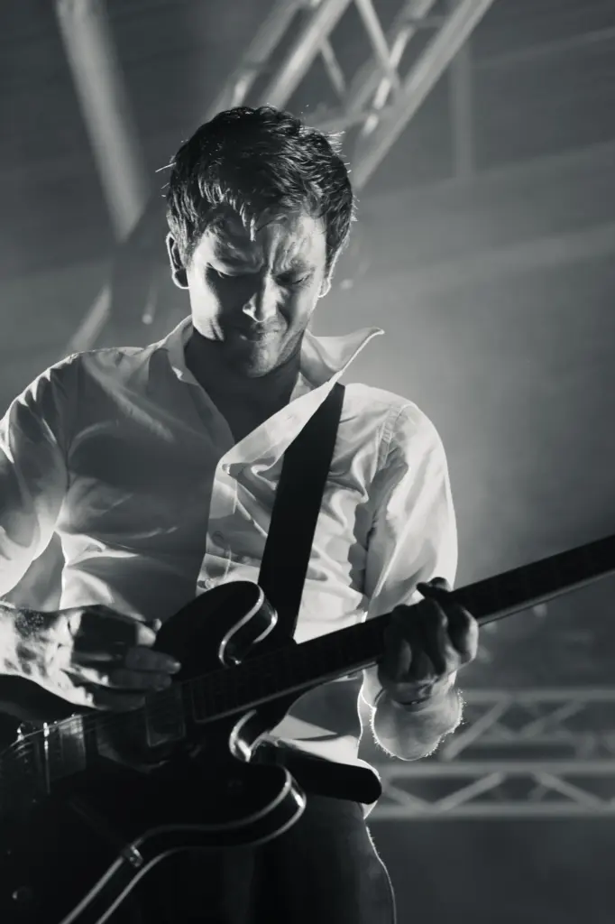 Musician playing a black electric guitar, wearing a white shirt, captured in dramatic stage lighting.