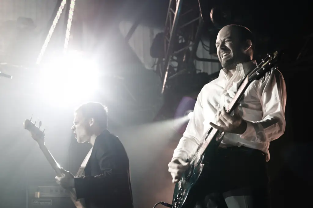 Two musicians performing on stage, one with a bass guitar, the other with an electric guitar, in a bright setting.