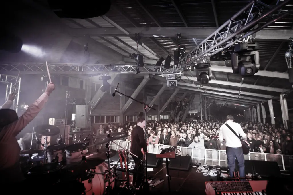 Band performing on stage with a large audience in a concert setting, illuminated by stage lights.