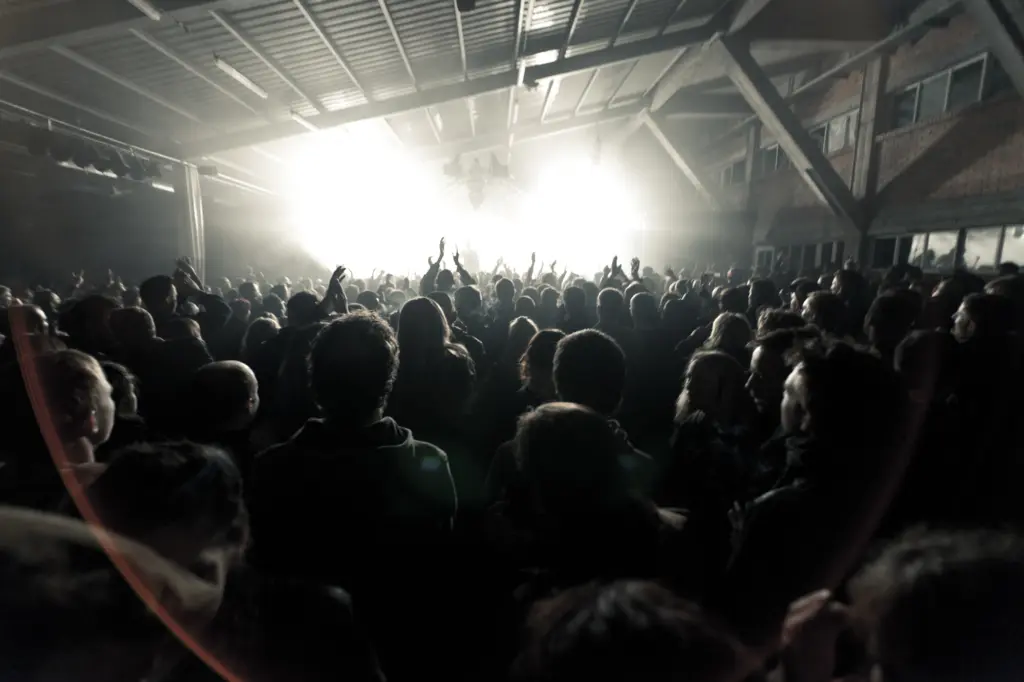 Audience with raised hands at a concert under bright stage lights in an indoor venue.