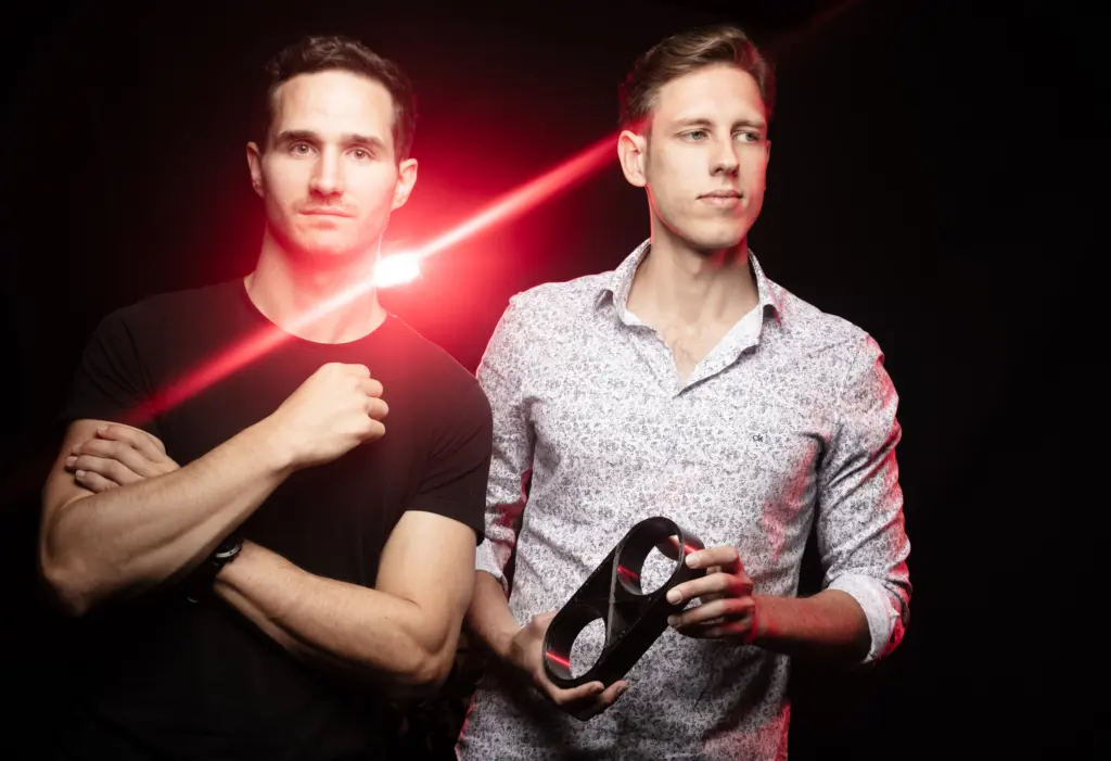 Two men standing together, one holding a mask, with a red light effect in the background.