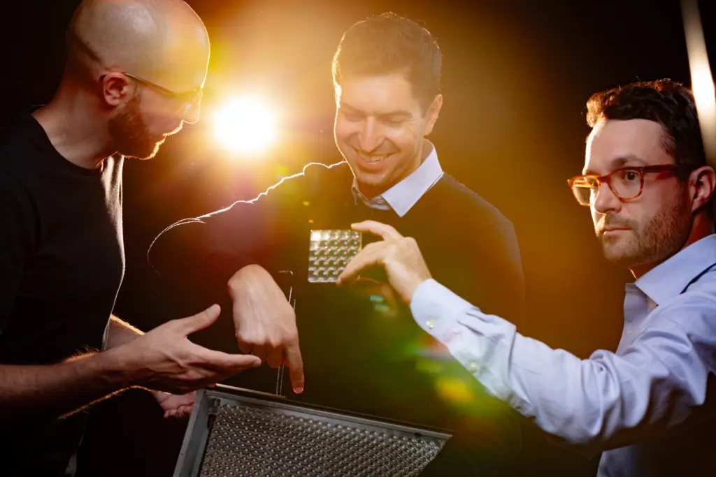 Three men interacting with a metallic light panel, wearing casual and formal attire, against a bright background.